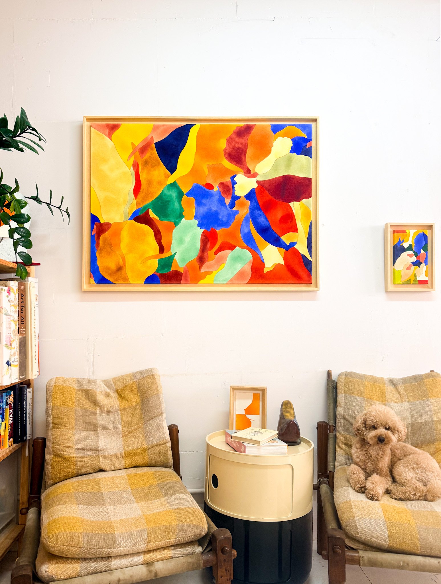 deux fauteuils en bois avec coussins en tissu à carreaux jaunes et beiges, un chien bouclé marron clair assis sur le fauteuil à droite, une petite table ronde beige avec des livres et des objets décoratifs, une huile sur toile abstraite avec des couleurs vives au mur, une étagère avec des livres et une plante à gauche.