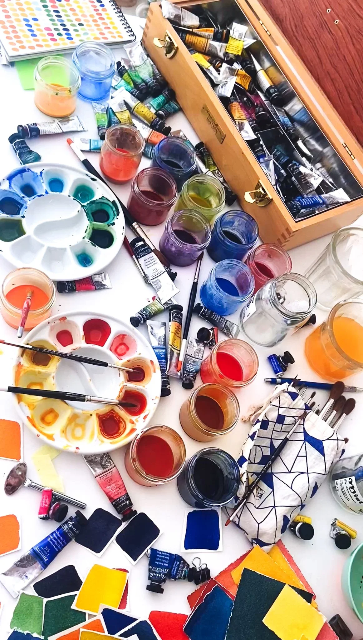 Espace de peinture avec des tubes de peinture acrylique, des pinceaux, une palette de couleurs, des verres contenant des peintures, des cotons-tiges, des échantillons de textures, et un coffret en bois remplie de tubes de peinture.