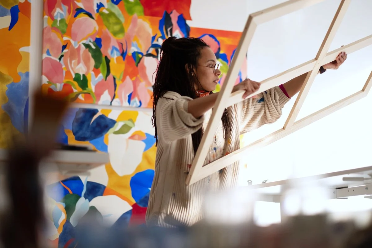 Une femme tenant une grande pièce de bois en forme de cadre dans un studio d'art, avec une peinture florale colorée en arrière-plan.