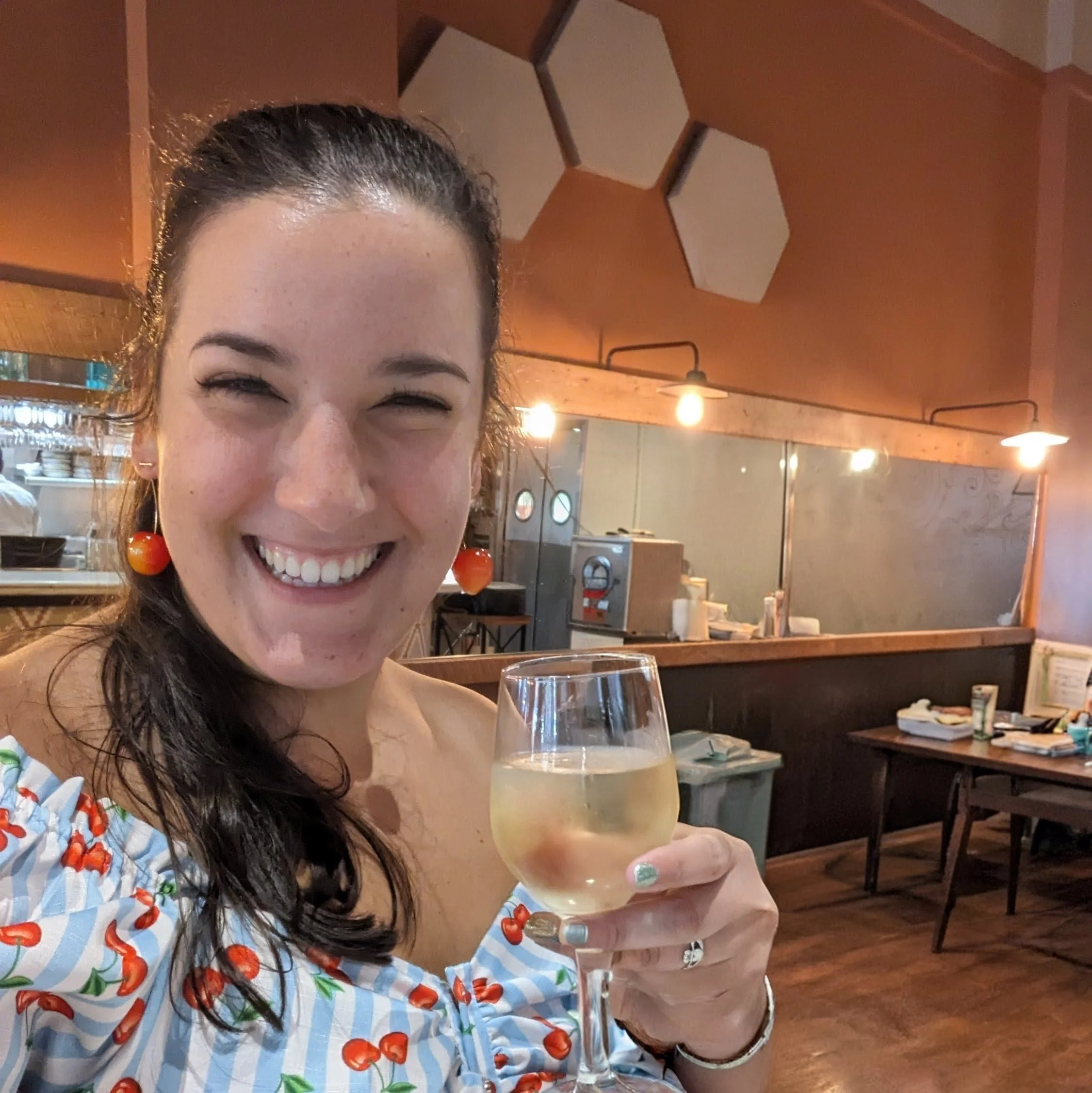 A woman with long dark curly hair smiling and holding a glass of champagne in a restaurant with warm lighting and orange walls.