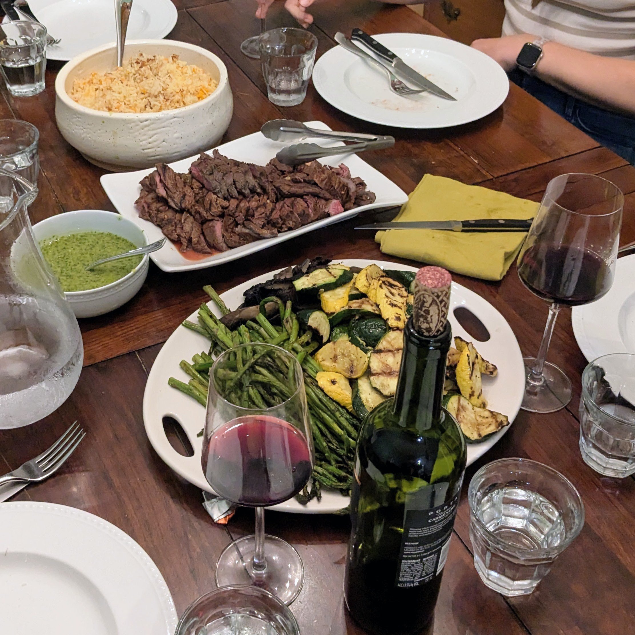A dinner table with plates of grilled meat, roasted vegetables, a bowl of rice, and a small bowl of green sauce. Several glasses of red wine and water are on the table. The table is made of wood, and there are utensils and napkins placed around.