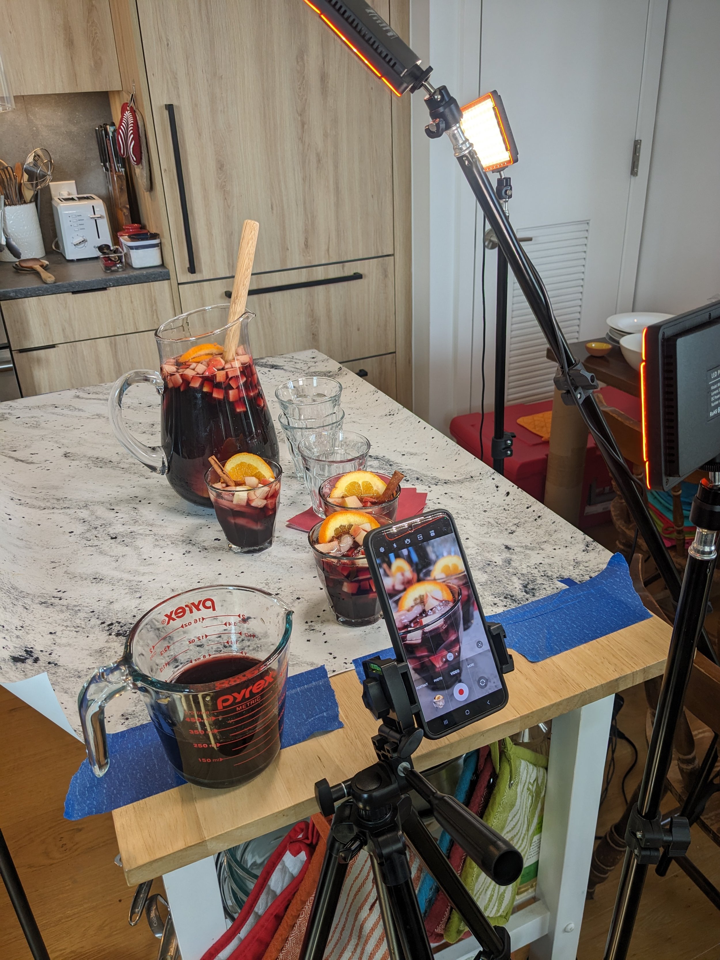 A kitchen countertop with a glass pitcher and matching glasses filled with a dark red beverage, garnished with orange slices, cinnamon sticks, and fruit pieces. A smartphone on a tripod captures the scene. There's a blue paper towel under the pitcher, and kitchen items like a toaster and utensils are in the background.