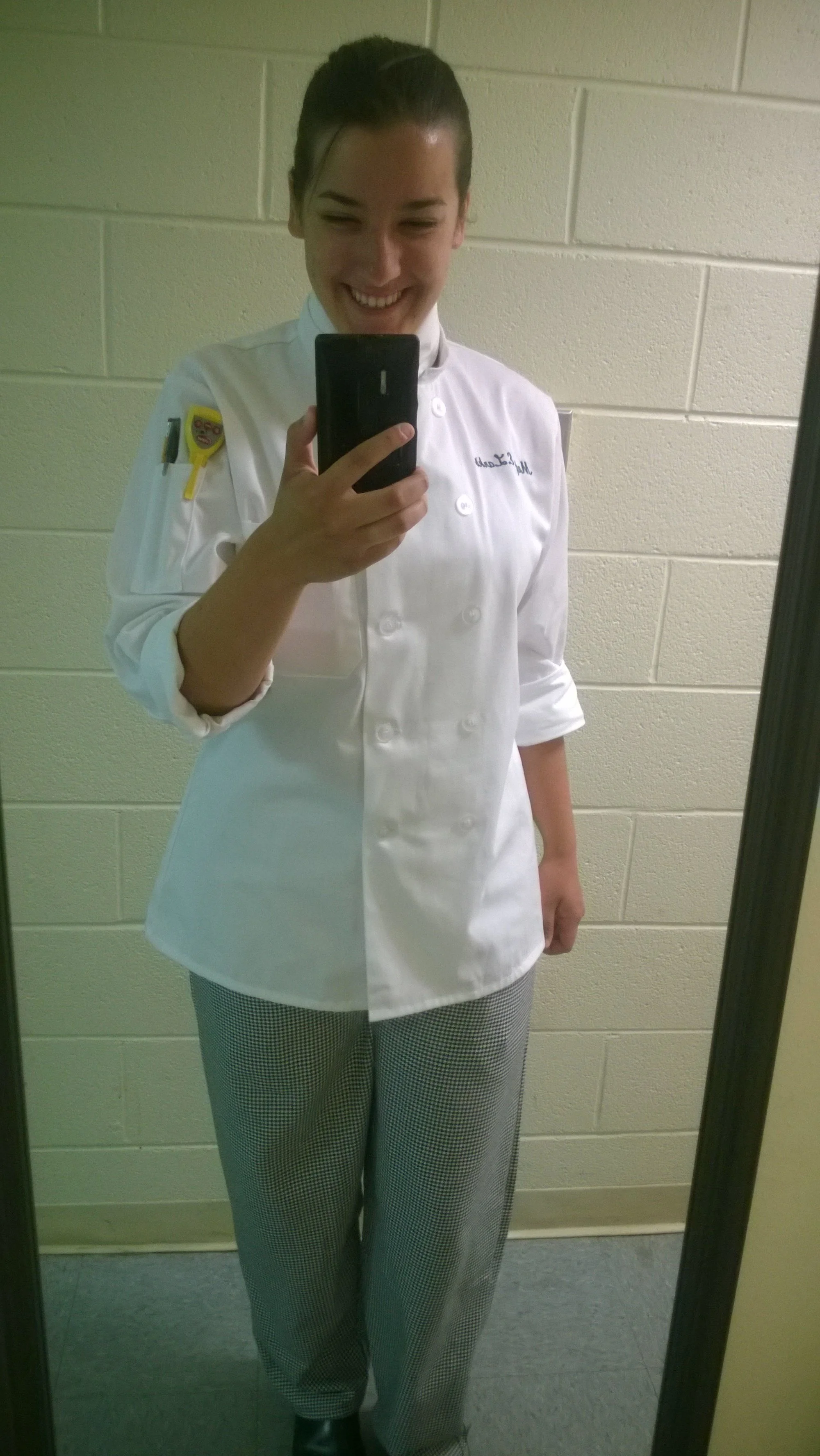 A person wearing a white chef's coat with a name embroidered on it, taking a selfie in front of a mirror. The person is smiling and holding a smartphone, with a wall and mirror frame visible in the background.