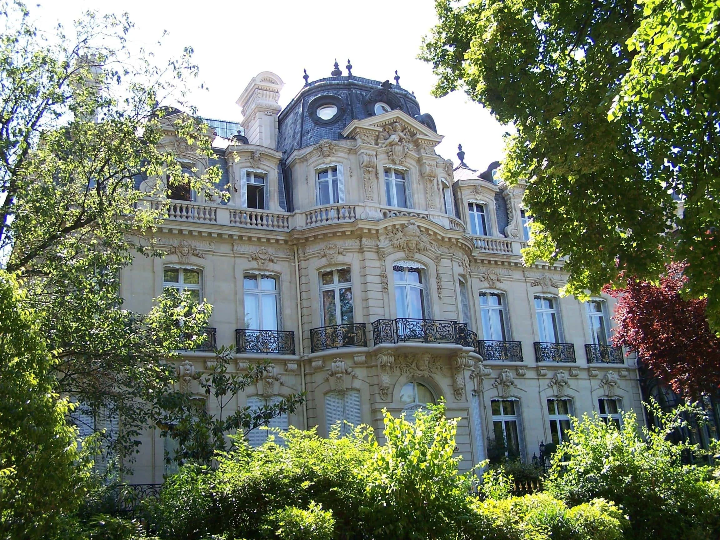 Façade d'un bâtiment historique de style haussmannien avec des fenêtres grandes et un toit en mansarde, entouré d'arbres verts évoquant un jardin ou un parc.