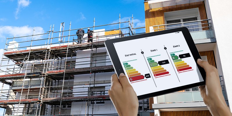 Une personne tient une tablette affichant un rapport énergétique, avec un bâtiment en construction en arrière-plan.