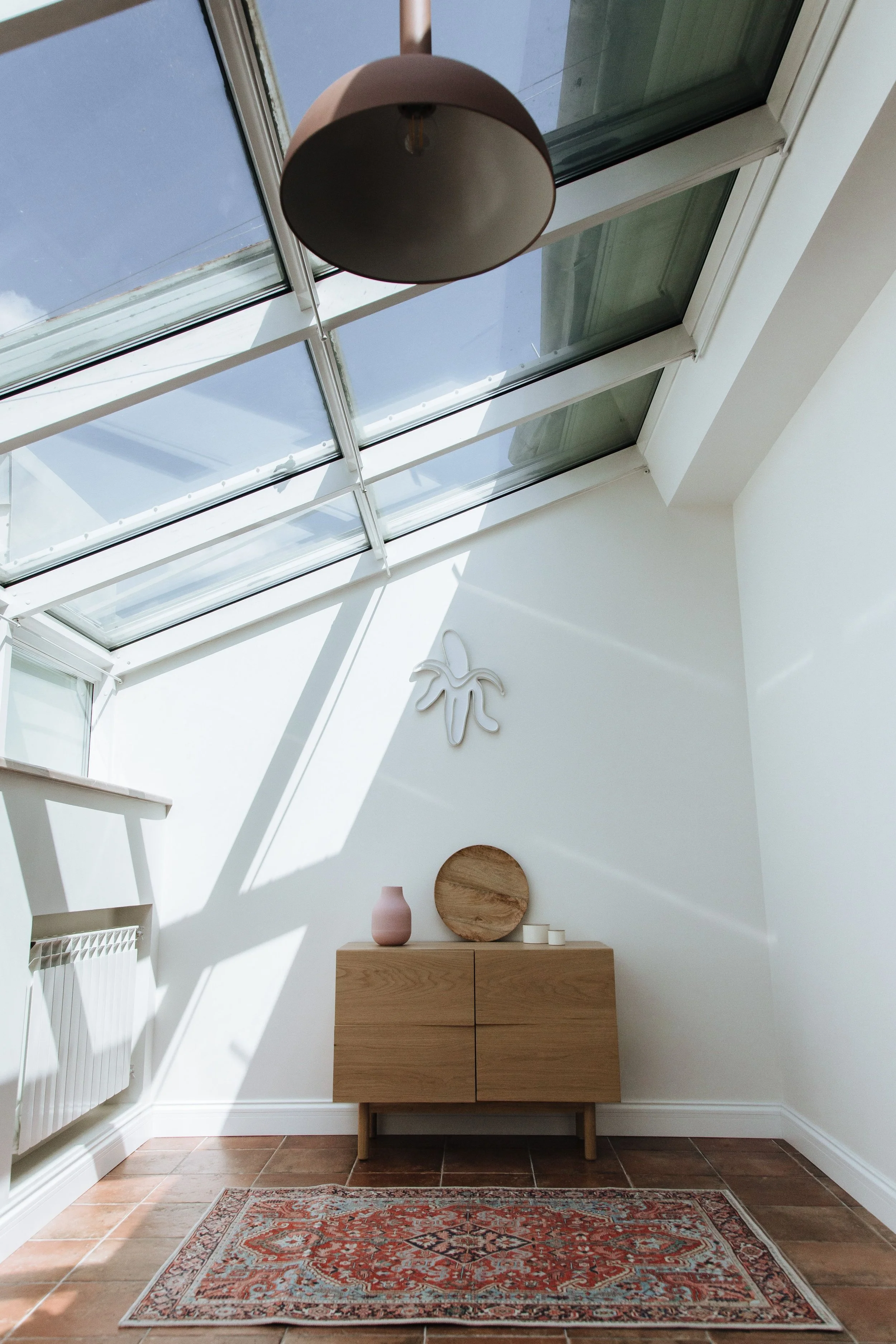 Un intérieur moderne avec un plafond incliné en verre laissant entrer la lumière naturelle, une commode en bois clair, un vase rose, un plateau en bois, et un tapis oriental sur un sol en carrelage de couleur terre cuite.