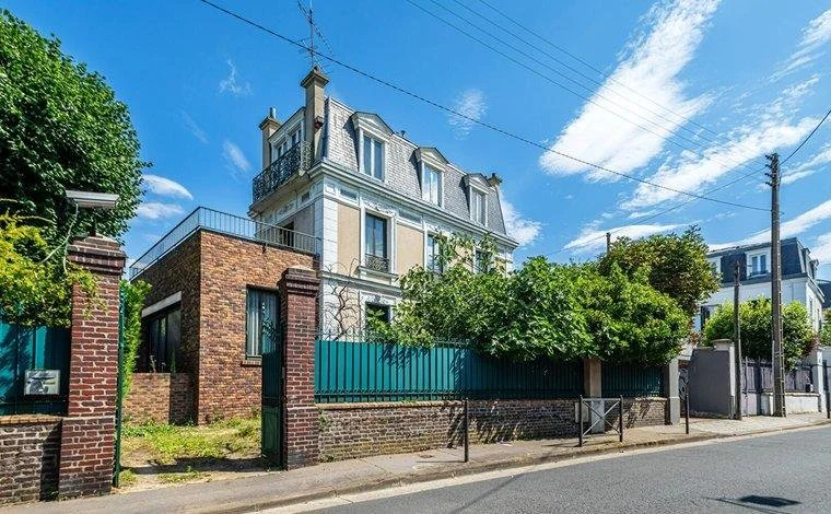 Une maison de style haussmannien avec un toit en mansarde, entourée d'un mur en brique et d'une clôture verte, située dans une rue résidentielle ensoleillée avec un ciel bleu.