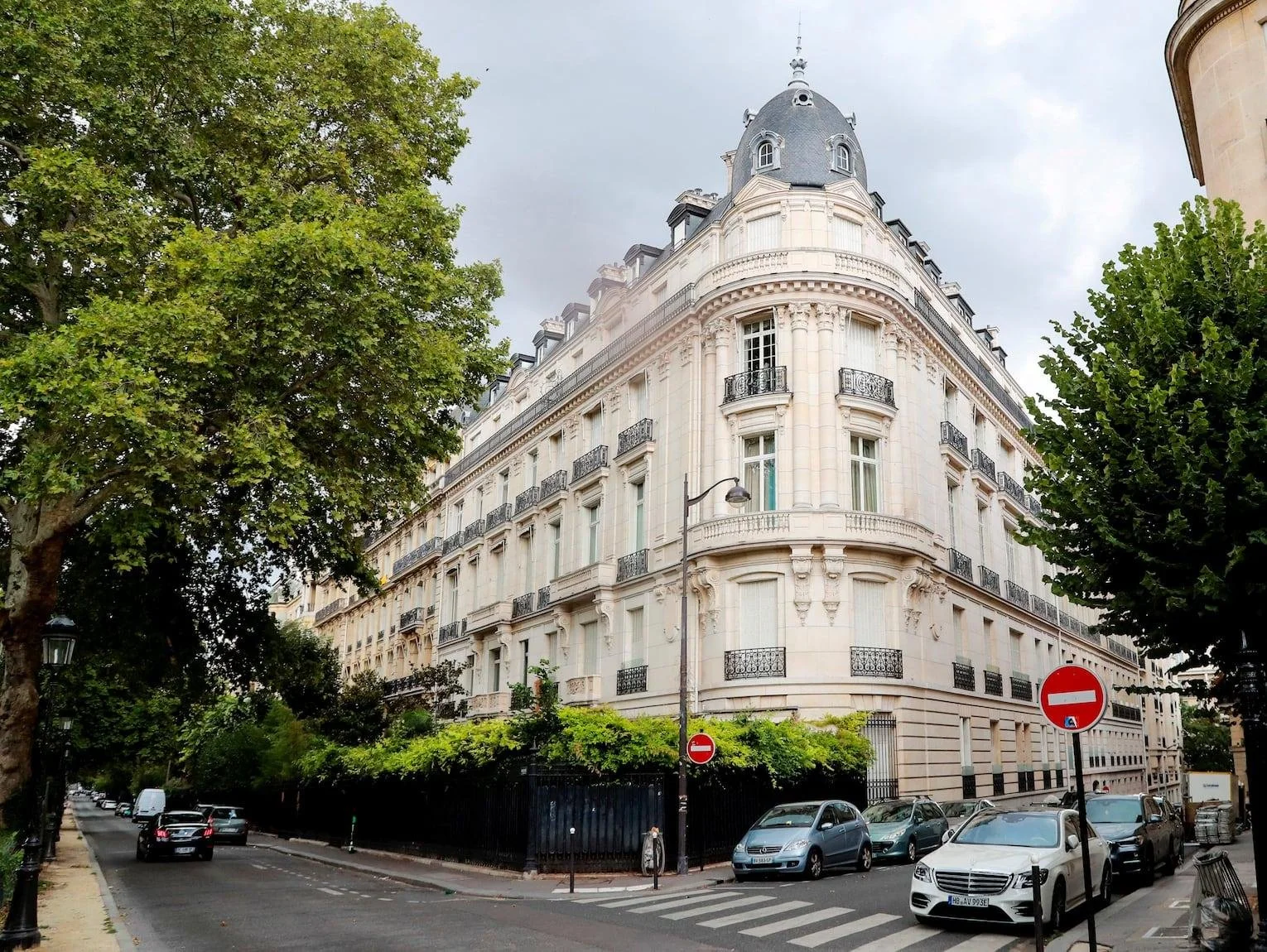 Bâtiment haussmannien en pierre blanche avec balcons en fer forgé, situé dans une rue bordée d'arbres, avec un ciel nuageux en arrière-plan.