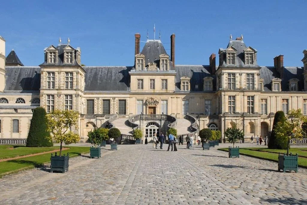 Un grand château avec une façade en pierre et de nombreuses fenêtres, entouré d'un jardin bien entretenu et de personnes qui se promènent.