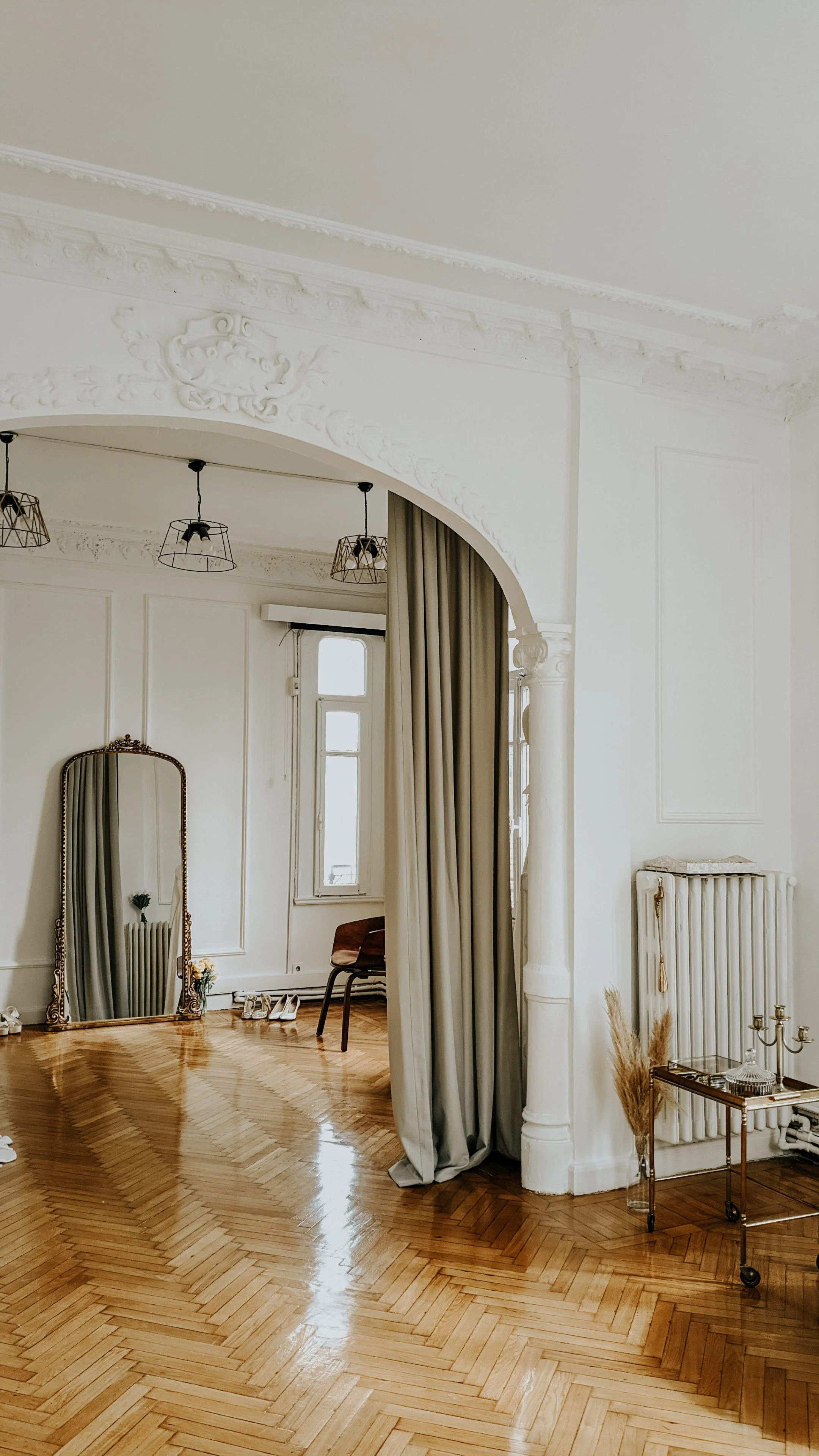 Intérieur d'une pièce élégante avec murs blanc cassé, parquet en bois clair, grands miroirs, rideaux gris, et meubles décoratifs en laiton et verre.