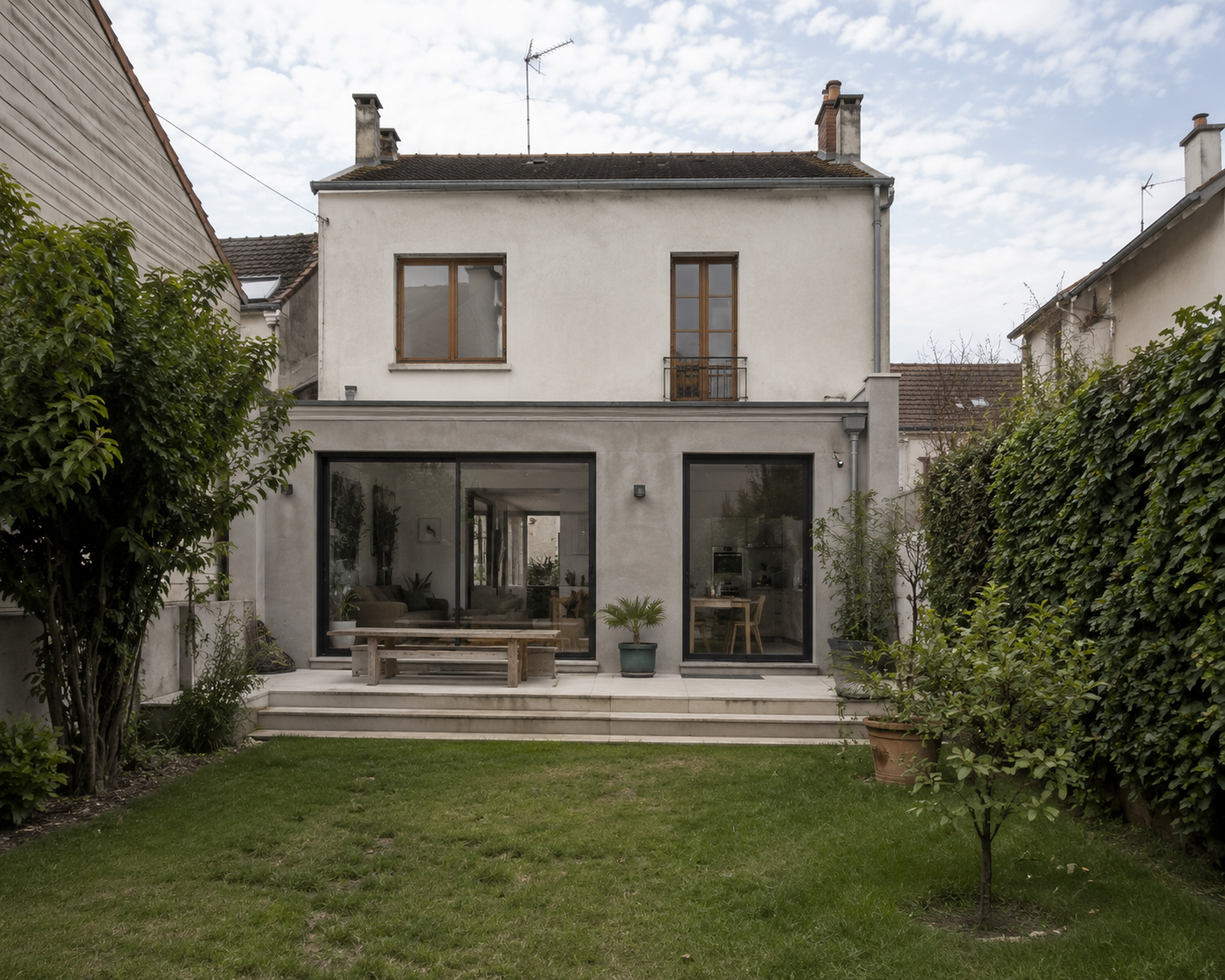 Vue d'une maison avec un jardin vert, terrasse et grandes fenêtres, sous un ciel partiellement nuageux.