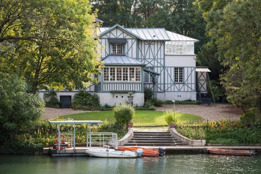 Une maison sur une colline bordée d'arbres, avec un perron, un jardin et un quai avec un bateau sur l'eau.