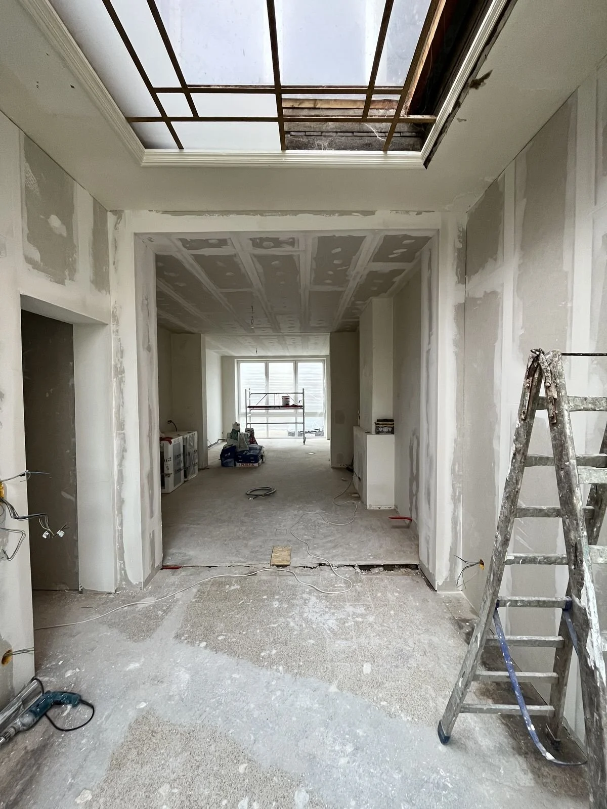 Intérieur en cours de rénovation d'un appartement, murs en plâtre avec travaux, escabeau en aluminium, plafond partiellement ouvert, fenêtres au fond, outils et matériaux de construction au sol.