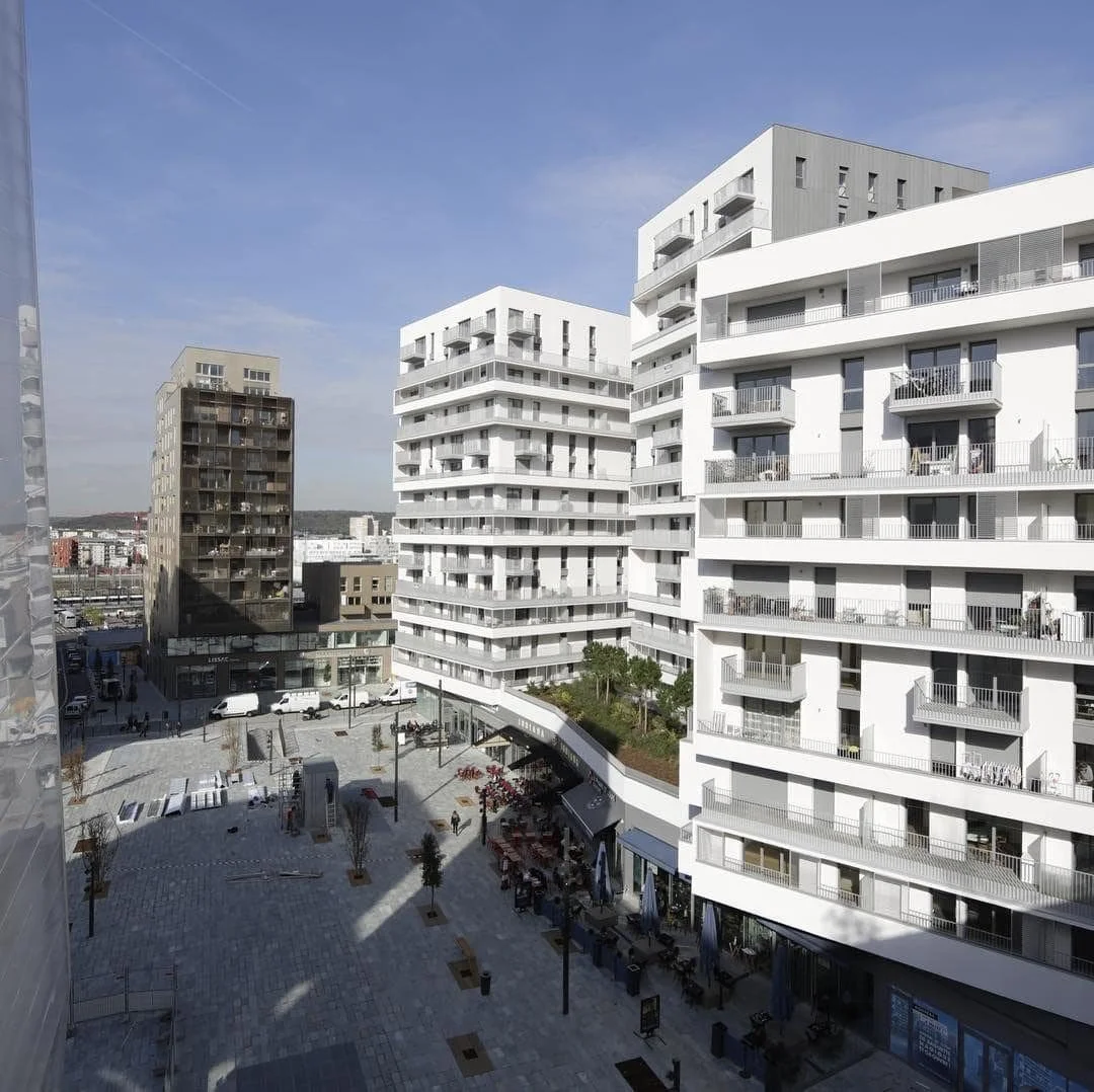 Vue d'un quartier avec plusieurs immeubles résidentiels modernes, un espace piétonnier avec des terrasses de cafés et des personnes. Ciel bleu avec quelques nuages.