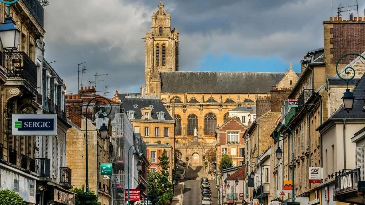 Vue d'une rue de ville en pente avec des bâtiments de style traditionnel, une église gothique en arrière-plan, et des nuages dans le ciel.