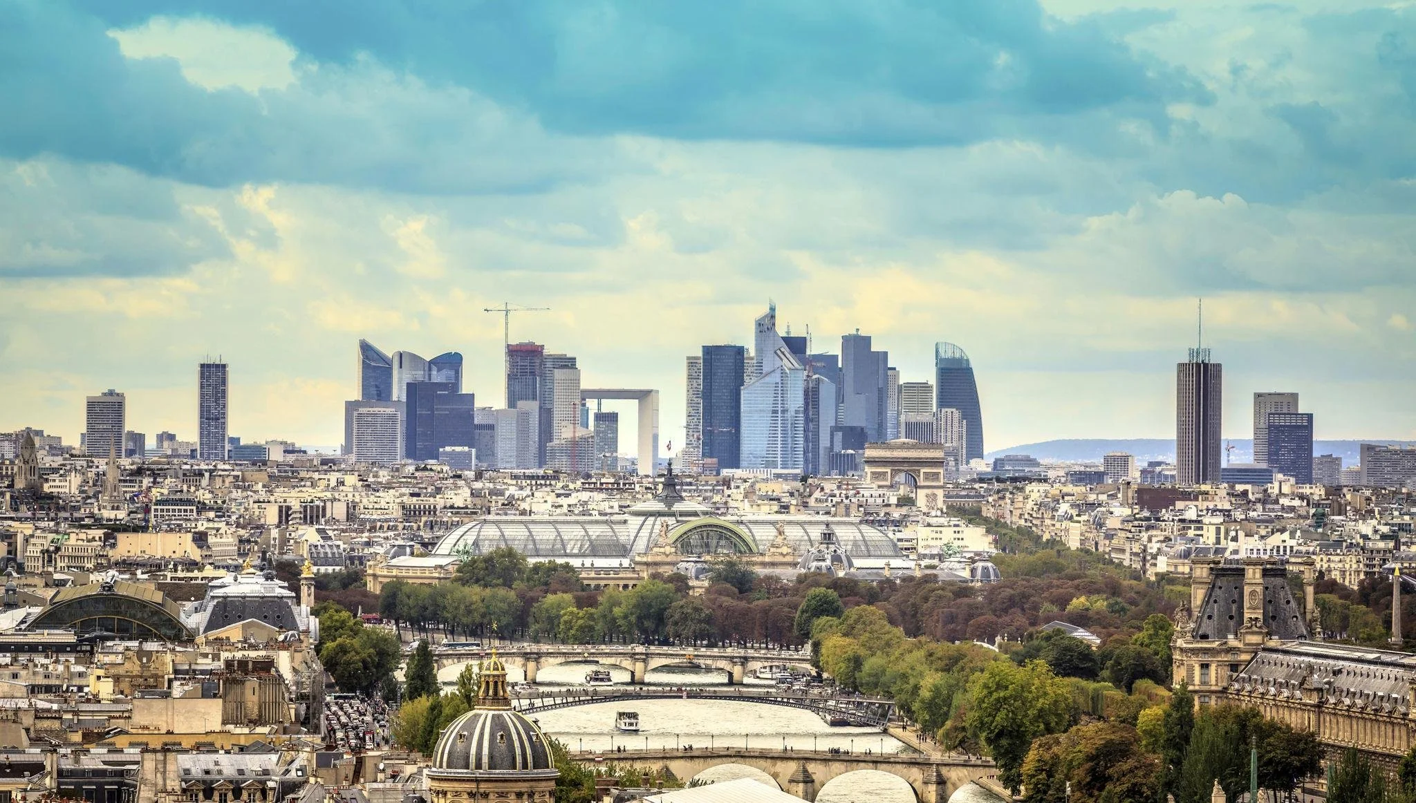 Vue panoramique de Paris avec la Seine, des bâtiments historiques en premier plan, et la skyline moderne avec de hauts gratte-ciel en arrière-plan sous un ciel nuageux.
