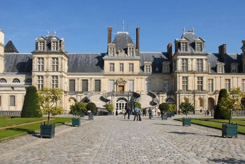 Un grand château avec une façade en pierre et de nombreuses fenêtres, entouré d'un jardin bien entretenu et de personnes qui se promènent.