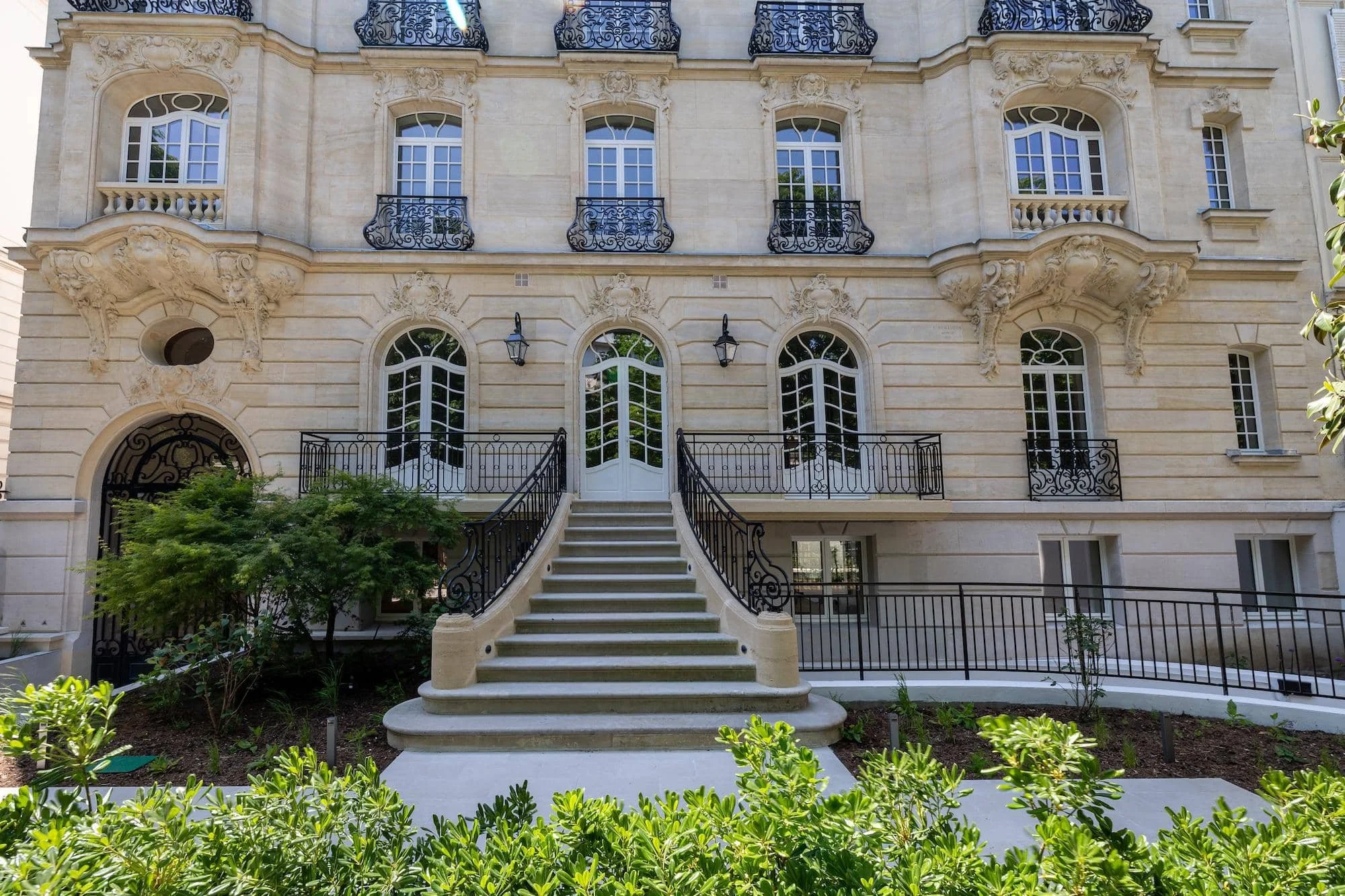 Façade d'un bâtiment élégant avec des fenêtres à arc, des balcons en fer forgé noir, un escalier en pierre menant à l'entrée principale, entourée de végétation urbaine