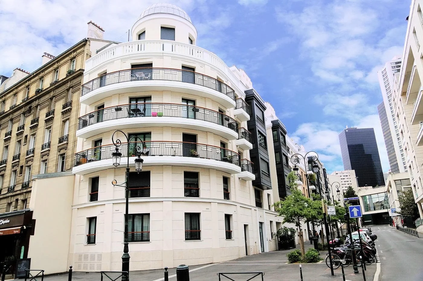Rues de Paris avec immeubles modernes et anciens, lampadaires en fer forgé, bicyclettes, une face d'immeuble blanc avec balcons noirs et une tour noire en arrière-plan, ciel partiellement nuageux.