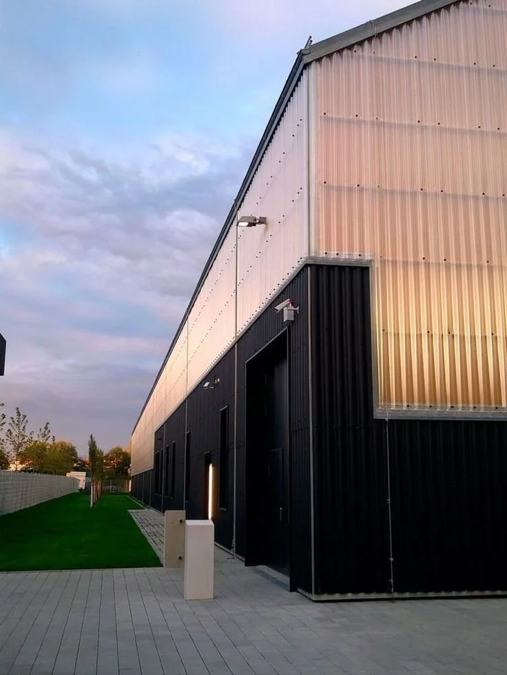 Une bâtiment moderne avec une façade en panneaux métalliques noirs et un toit incliné, sous un ciel partiellement nuageux.