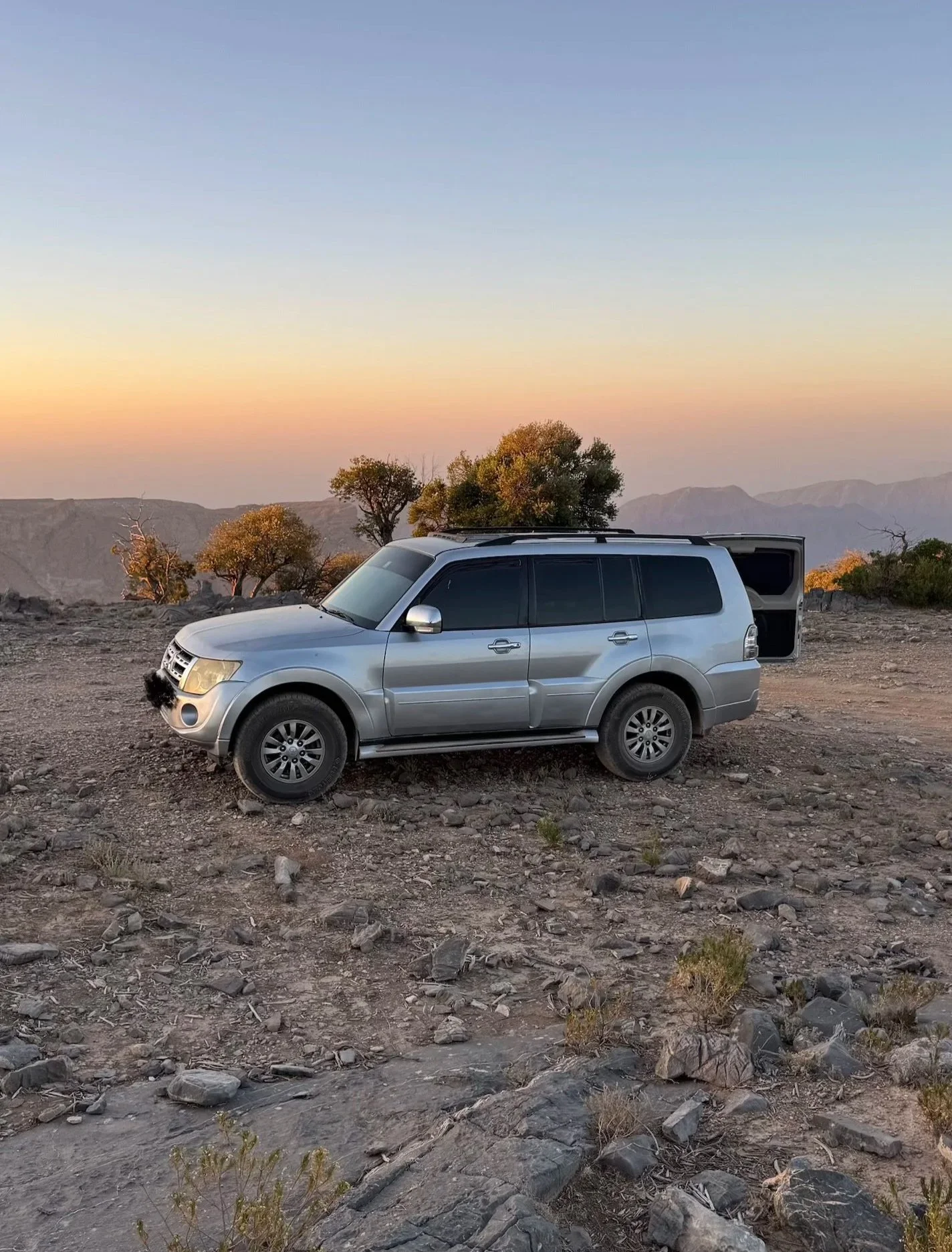 Ein silberner SUV steht auf einer felsigen, trockenen Landschaft bei Sonnenuntergang, im Hintergrund sind Bäume und Berge sichtbar.