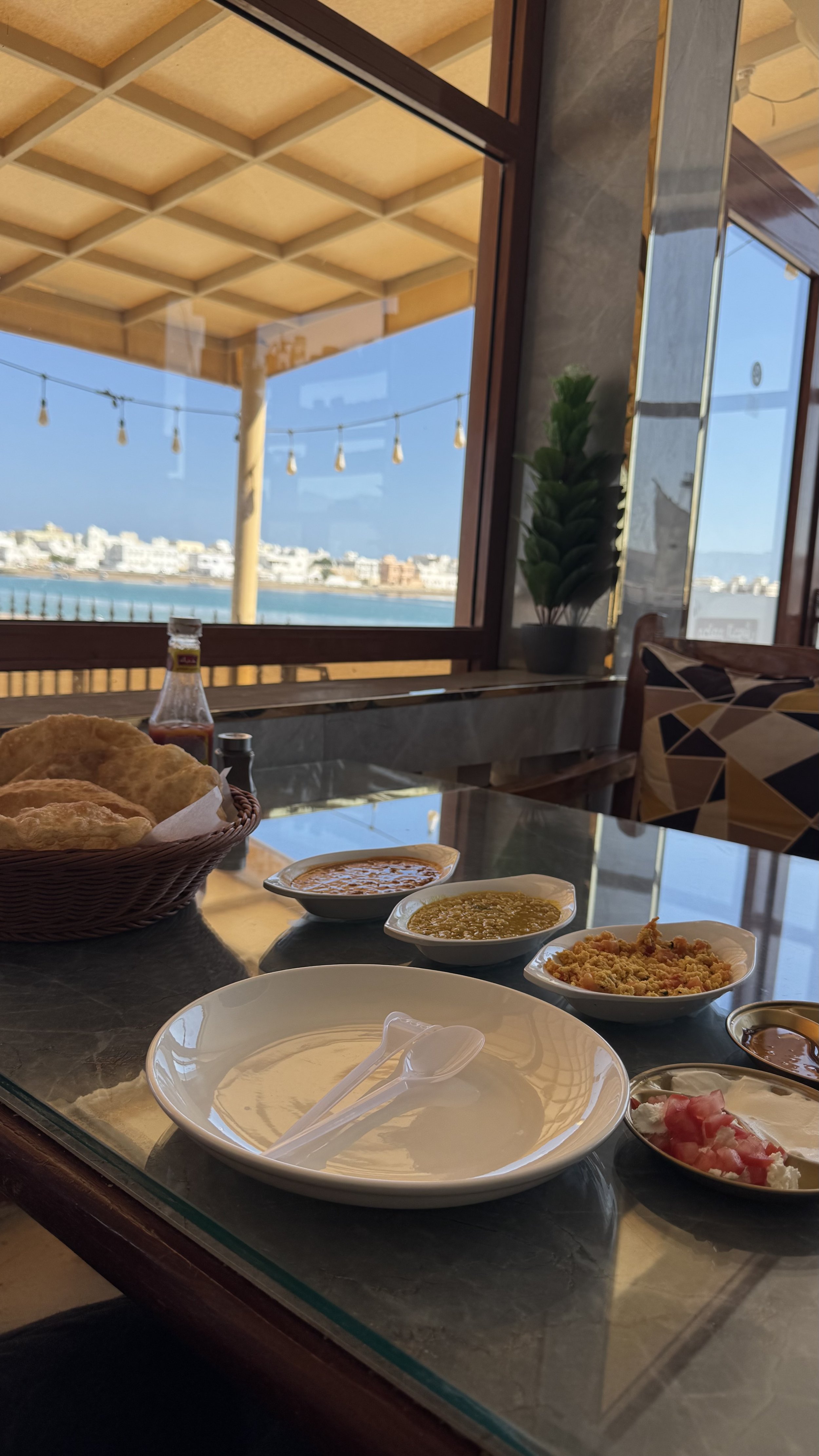 Ein Esstisch mit verschiedenen indischen Gerichten, darunter Chutneys, Papadam, Reis und Currys, vor einem Fenster mit Blick auf den Strand und eine Strandpromenade.