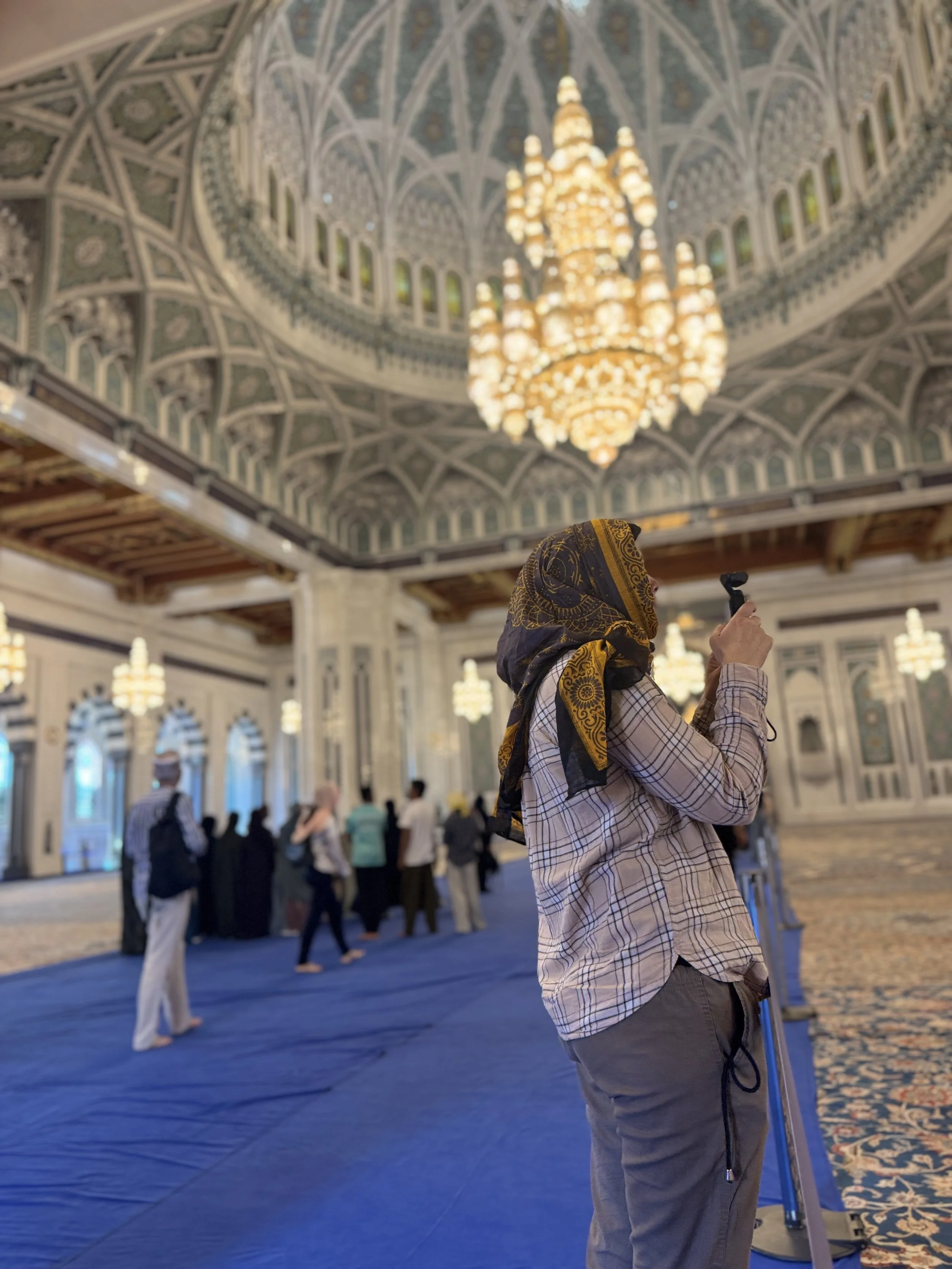Eine Person mit brauner, gelb-gestreifter Kopfbedeckung, grauem Hemd und grauen Hosen steht in einer prächtigen Halle und fotografiert mit einer Kamera. Im Hintergrund sind mehrere andere Personen zu sehen, sowie große, prachtvolle Kronleuchter und eine kunstvoll verzierte Decke im weißen und goldenen Stil.