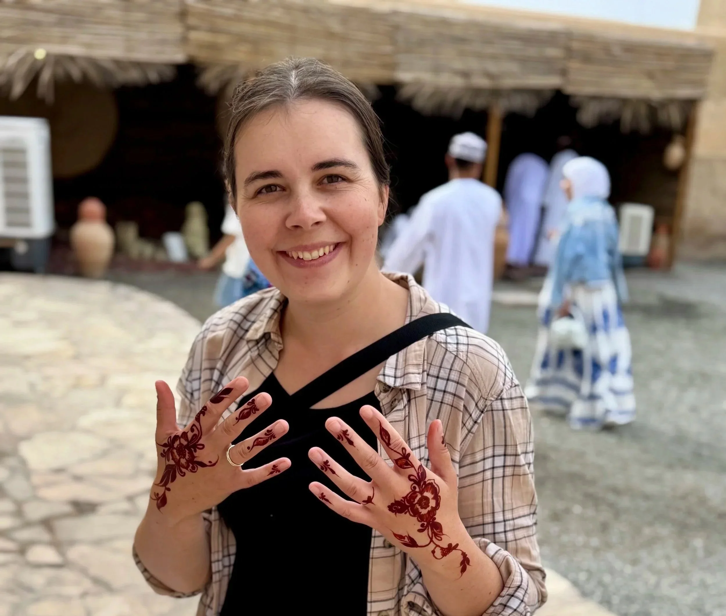 Junge Frau mit Henna-Handen vor einer Holzhütte, lachend, im Freien.