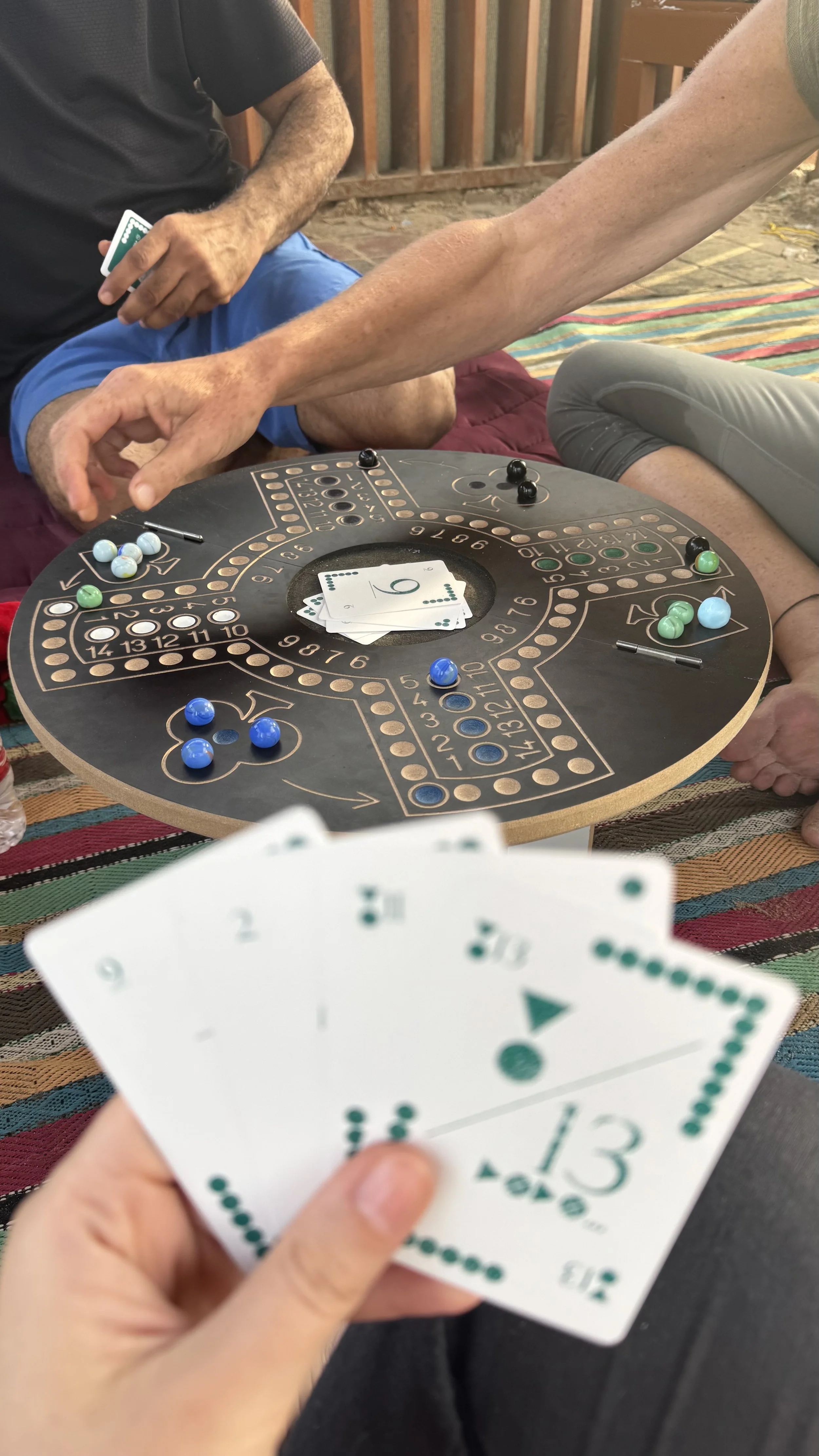 Menschen spielen ein Brettspiel im Freien. Im Vordergrund hält jemand Karten, im Hintergrund befindet sich eine runde Spielplatte mit Spielsteinen und Karten. Es ist ein geselliger Moment.