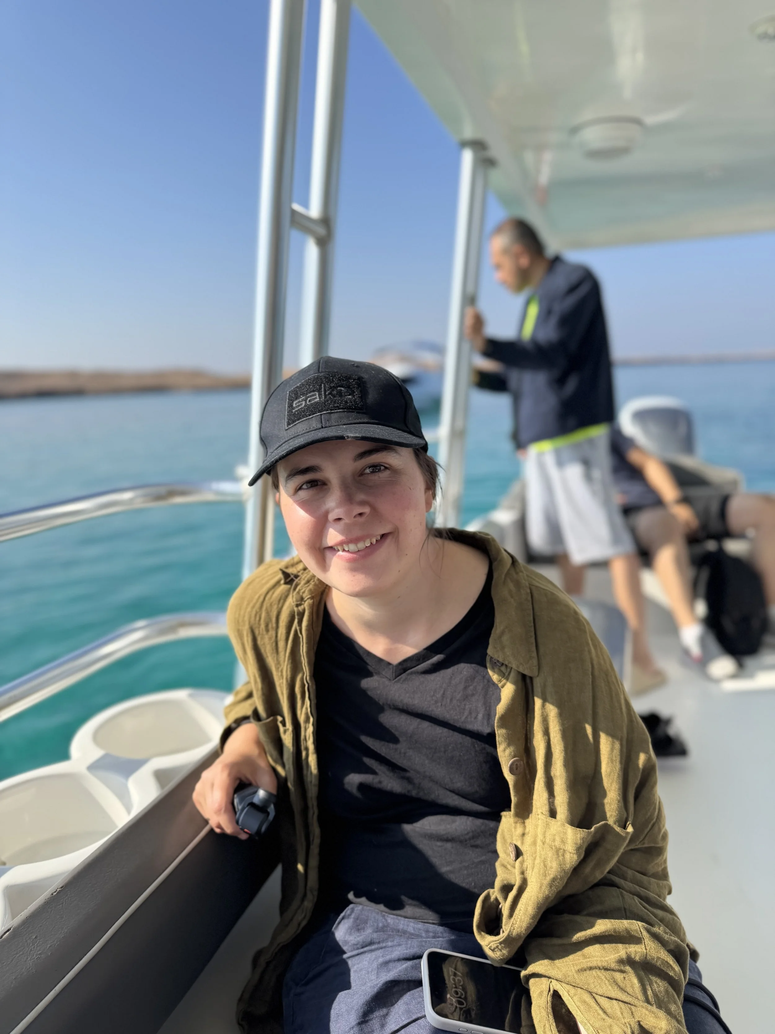 Junge Person mit schwarzer Cap und brauner Jacke sitzt auf einem Boot, lachend, während eine andere Person im Hintergrund liest, mit Blick auf das Wasser und den Himmel.