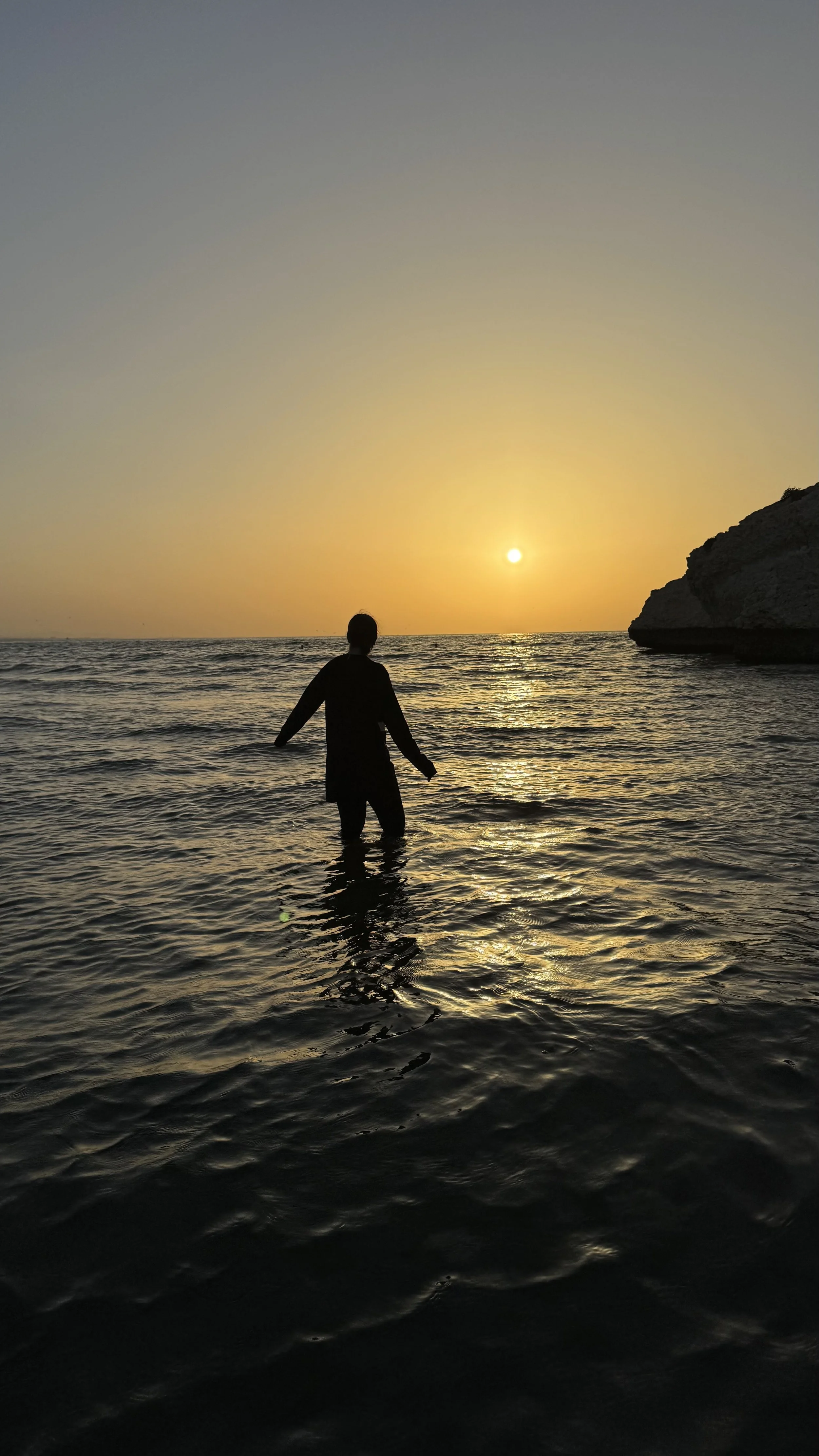 Silhouette einer Person im Wasser während eines Sonnenuntergangs am Strand mit Felsen im Hintergrund.