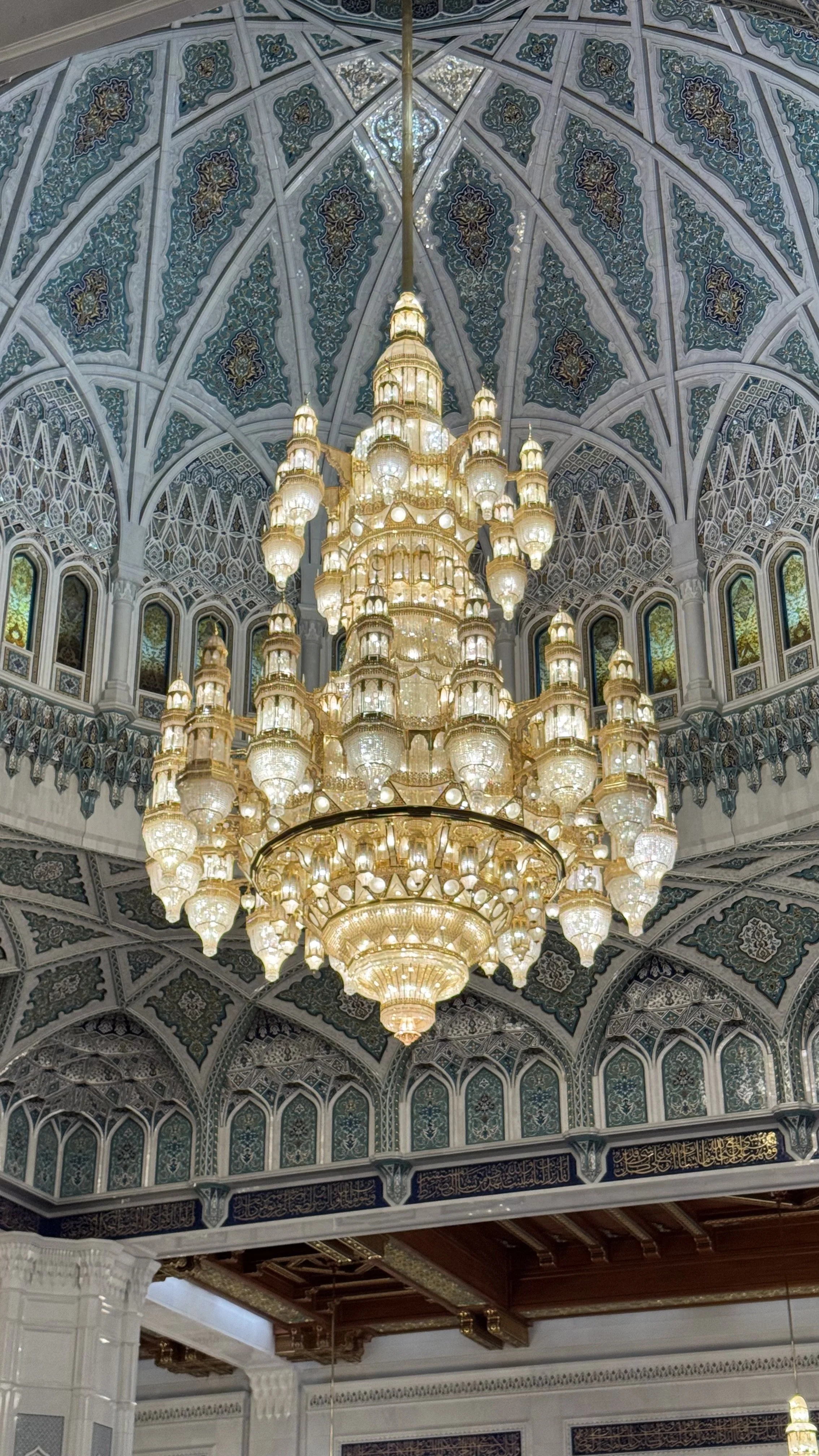 Ein großer goldener Kronleuchter mit vielen kleinen Lichtern hängt von der Decke in einem prunkvollen Raum mit eleganter Dekoration und kunstvollen Mustern.