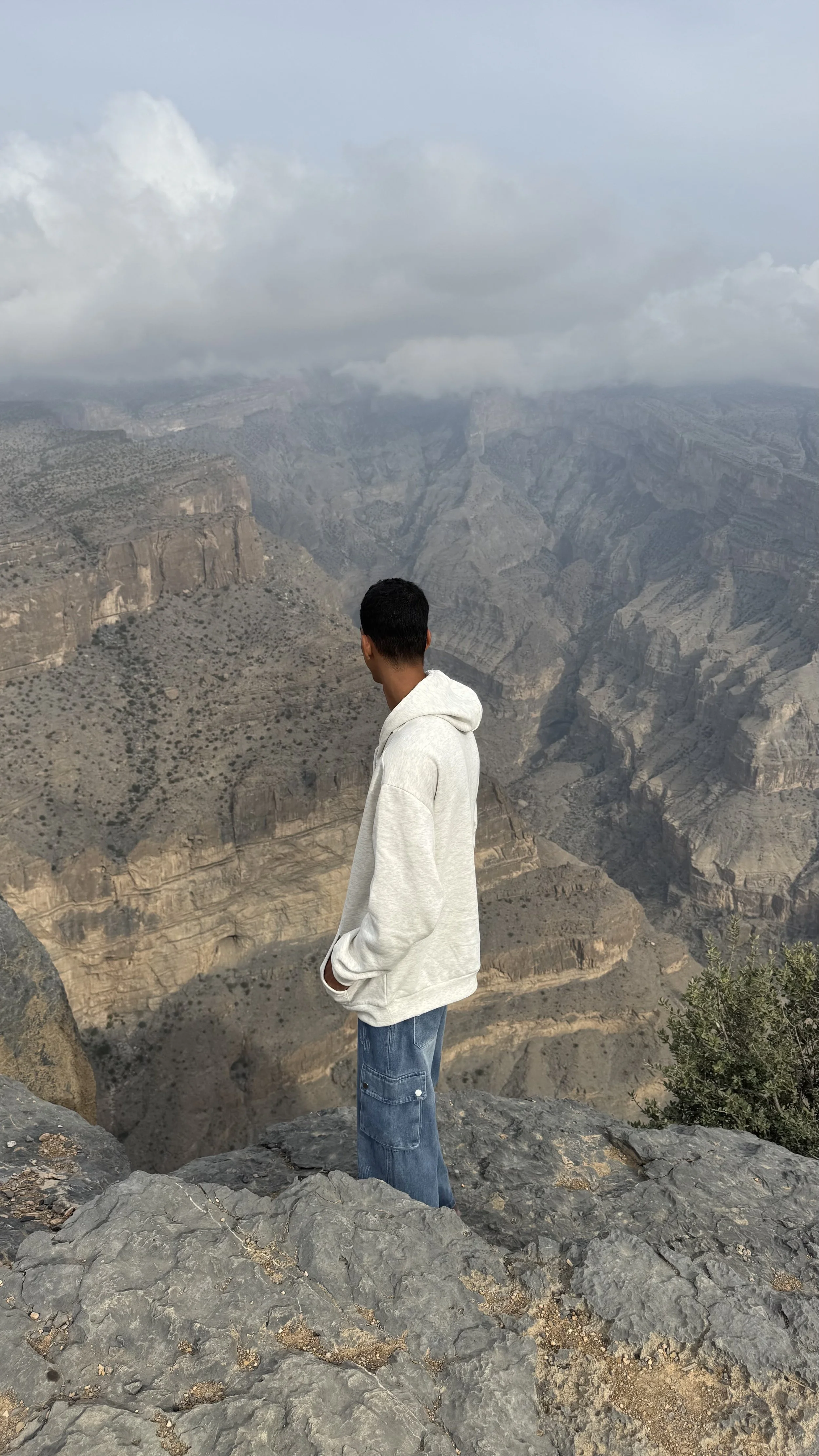 Junge Mann in weißem Hoodie und blauer Jeans steht auf einem Felsen im "Grand Canyon des Omans", schaut in die Ferne, mit Wolken am Himmel und den tiefen Schluchten des Canyons im Hintergrund.