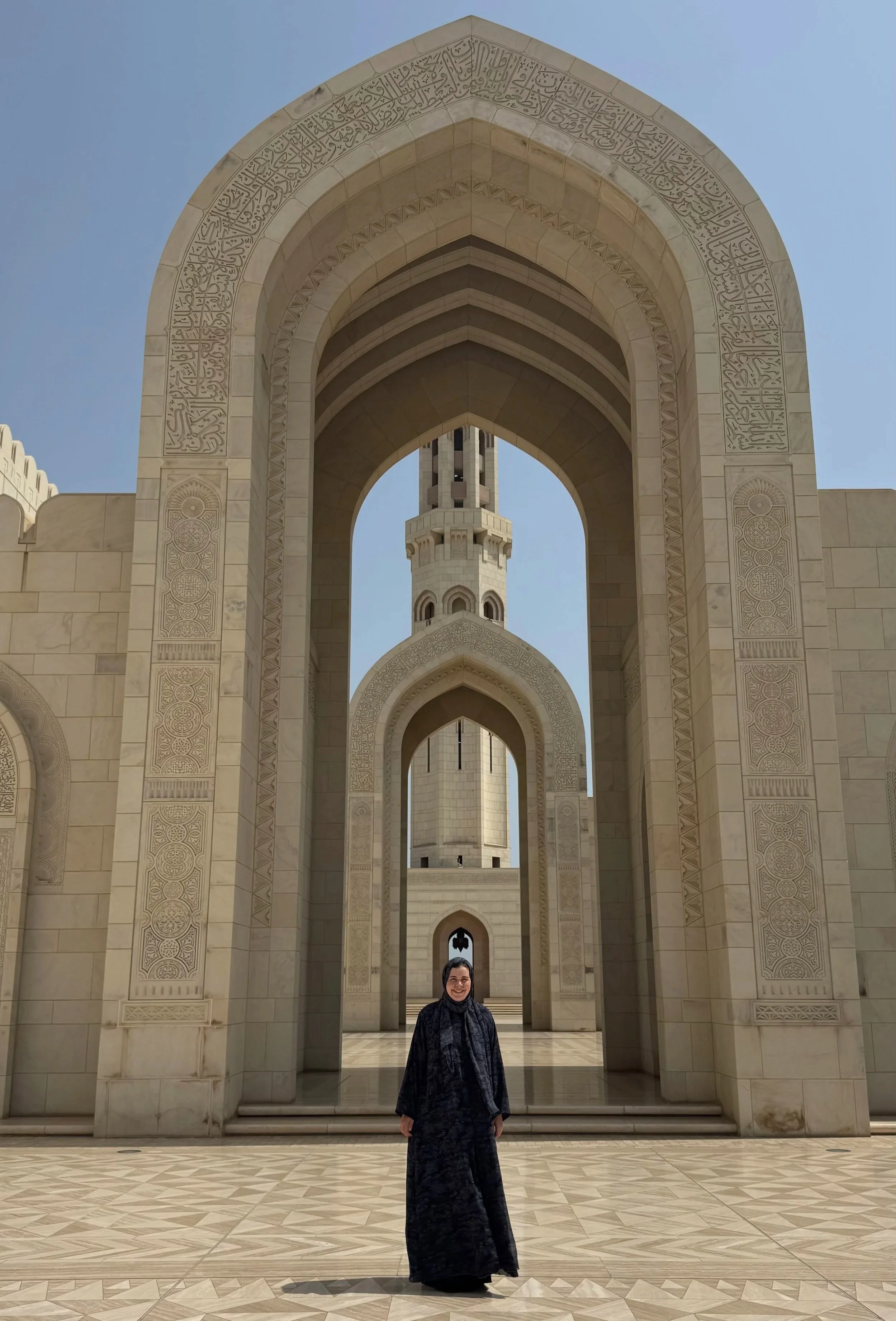 Eine Frau in schwarzem Kleid und Kopftuch steht vor einer großen islamischen Moschee mit hohen Torbögen und einer Minarett im Hintergrund, aus beige Marmor gebaut, bei sonnigem Himmel.