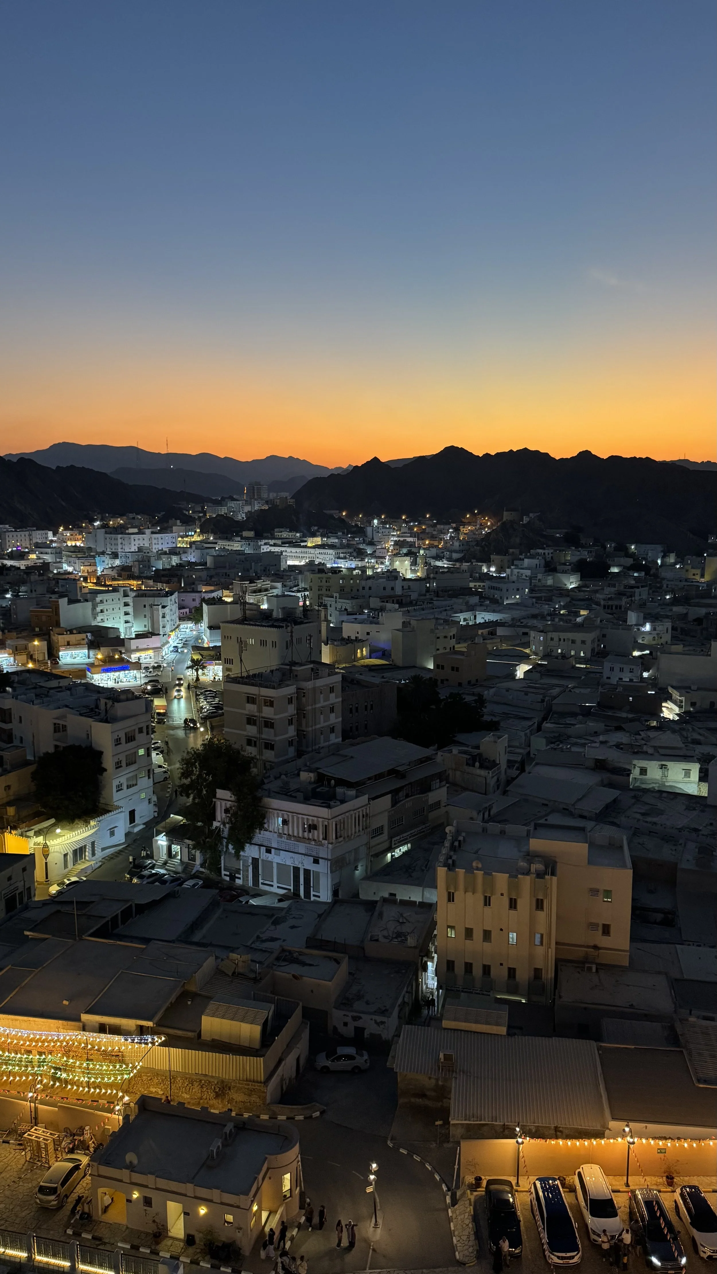 Städtische Skyline bei Sonnenuntergang mit Bergen im Hintergrund, beleuchtete Straßen und Gebäude im Vordergrund.
