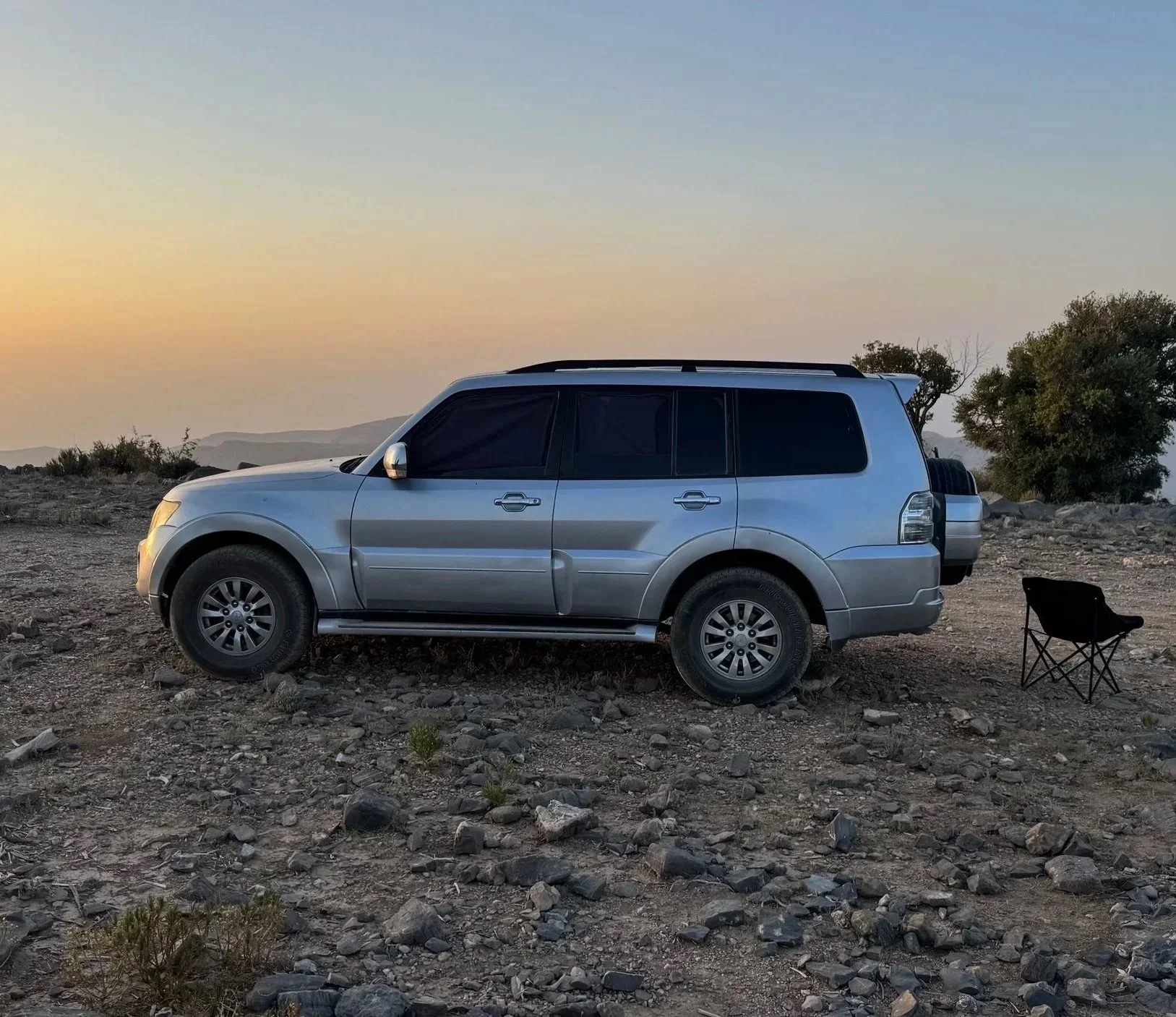 Ein silberner SUV steht auf einer felsigen, trockenen Landschaft bei Sonnenuntergang, mit einem Klappstuhl daneben.