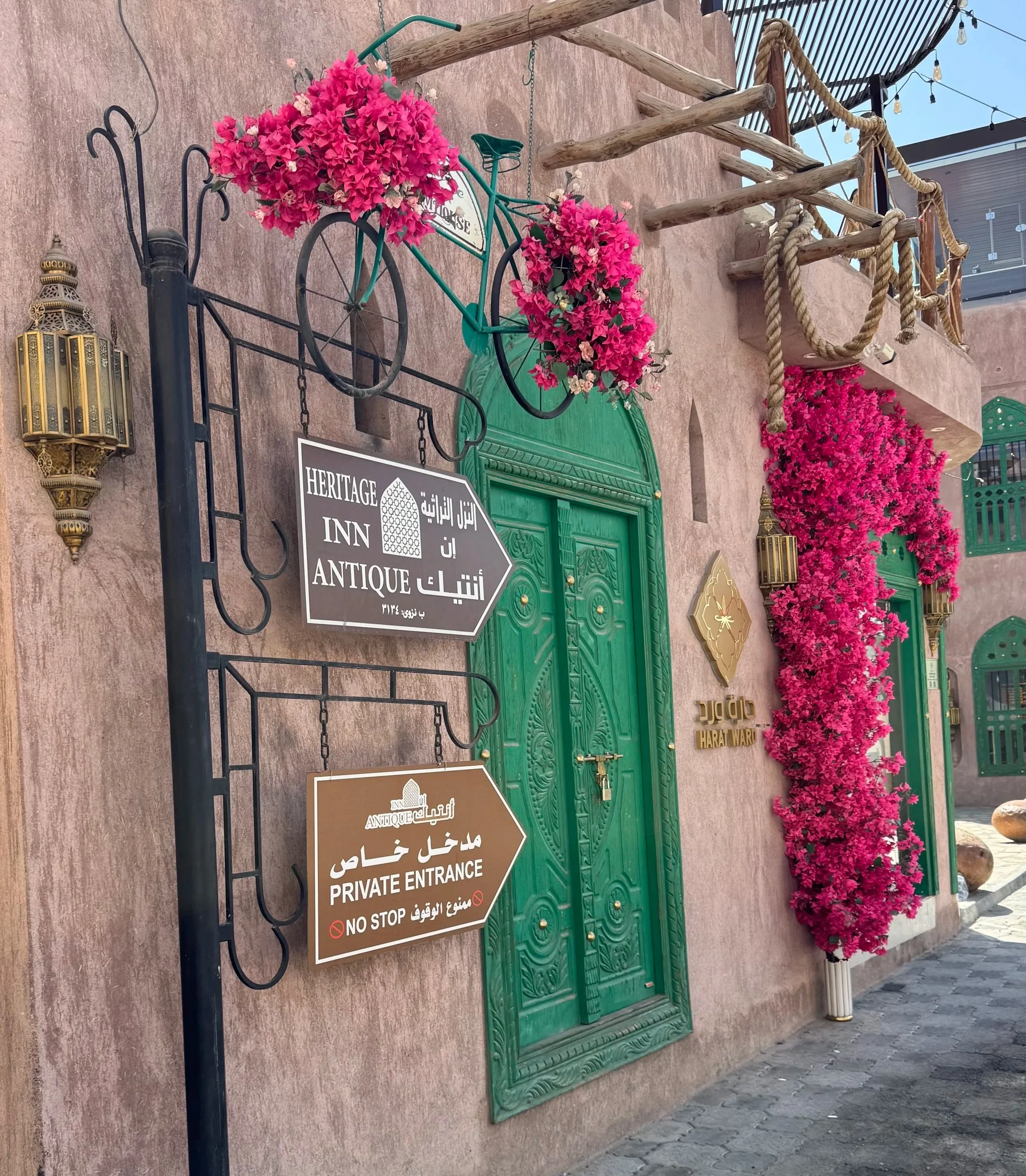 Ein grüner antiker Türrahmen mit einem schwarze. Fahrrad, das mit Pinken und Weißen Blumen dekoriert ist, hängt an einer Wand. Es gibt Schilder mit mehrsprachigen Hinweisen auf der Wand, darunter auf Englisch, Arabisch und Deutsch, die den Eingang als privat und antik kennzeichnen, mit Verboten für Stopp und Nicht-Stopp.