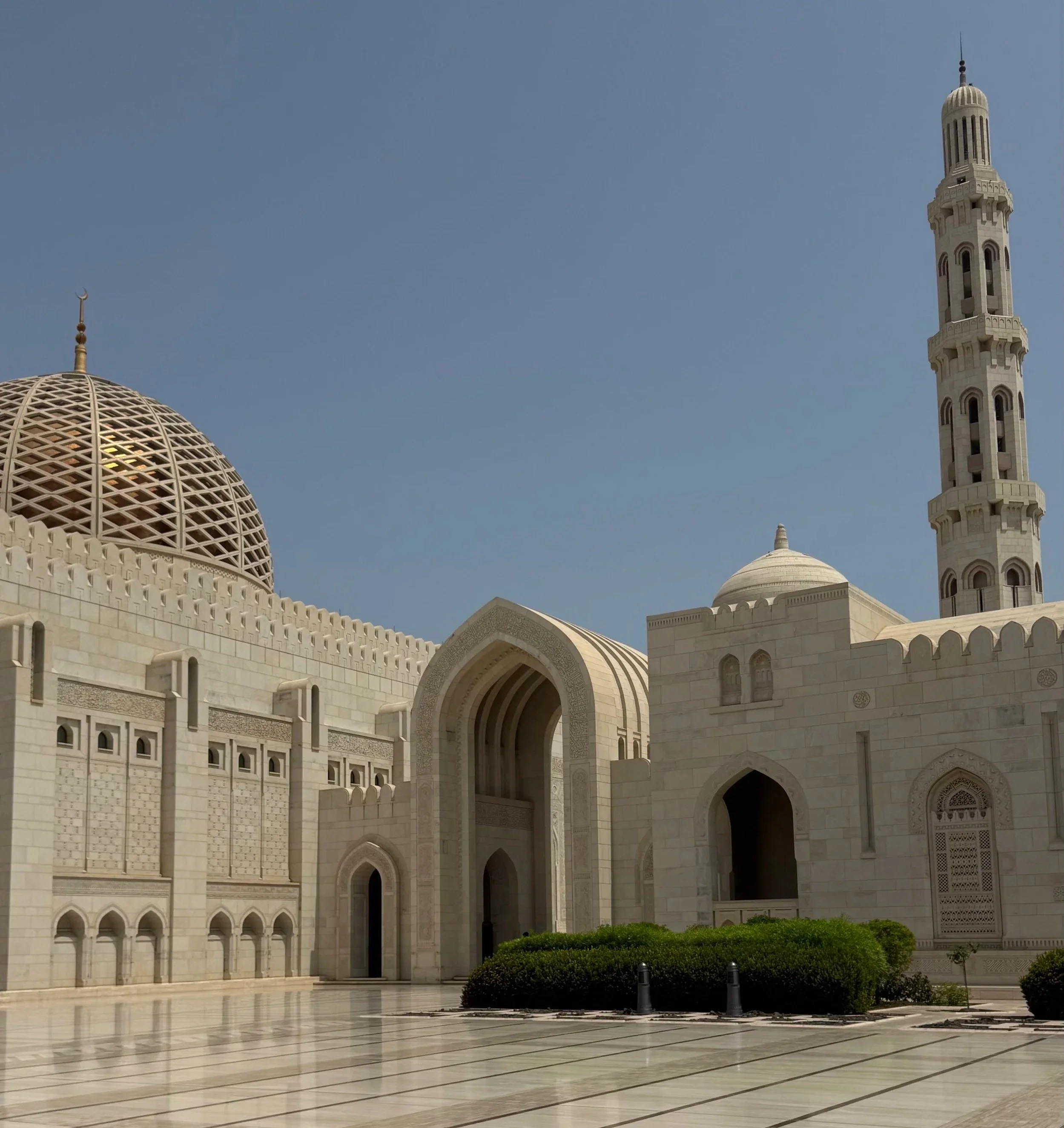 Ein prächtiges weißes Moschee-Gebäude mit Kuppeln und Minaretten vor blauem Himmel, umgeben von einem gepflasterten Vorplatz und kleinen Grünflächen.
