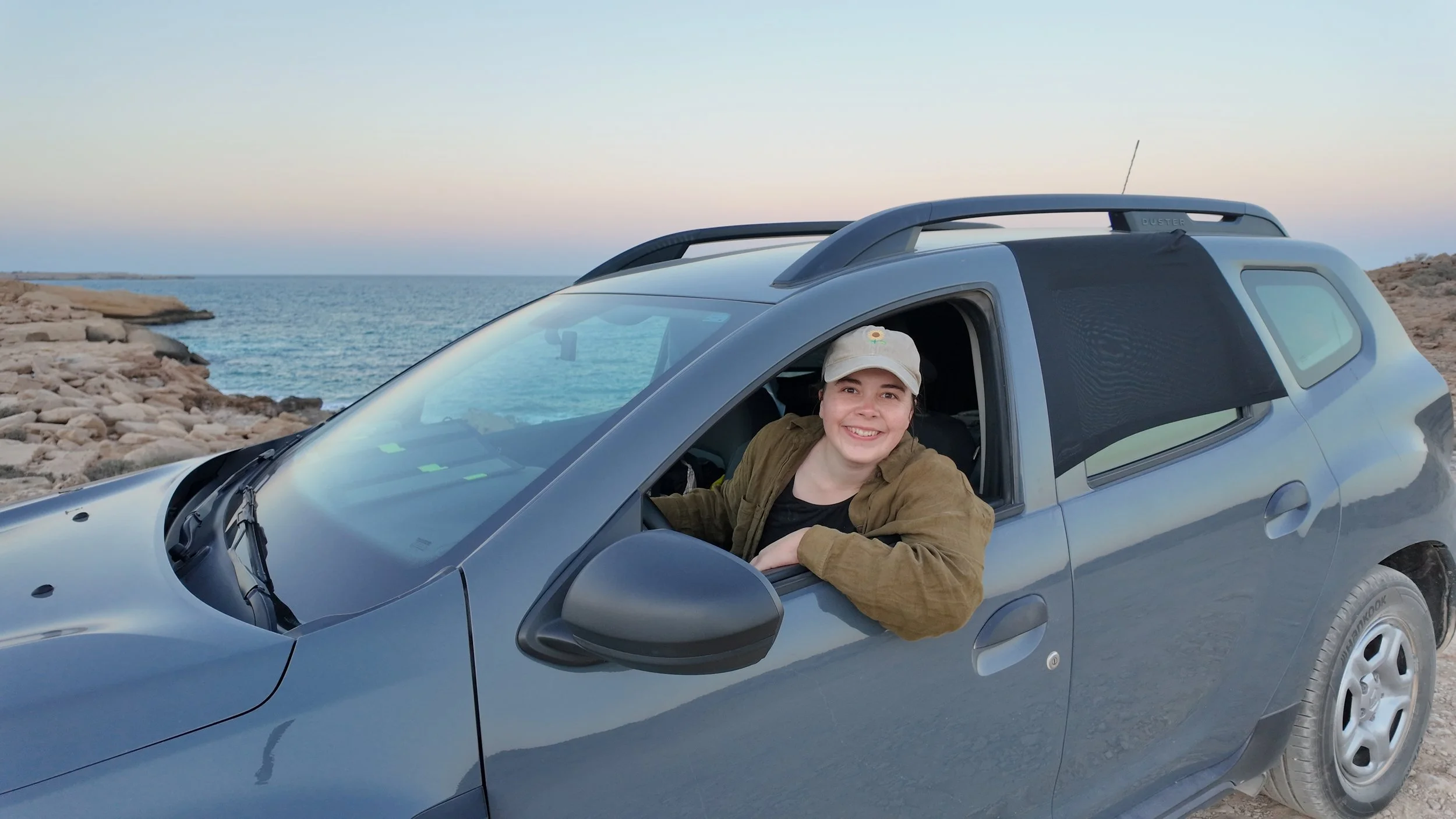 Lächelnde junge Frau im Auto am Meer, trägt Hut, das Auto ist auf einer felsigen Küstenstraße geparkt, Sonnenuntergang, ruhige Atmosphäre.