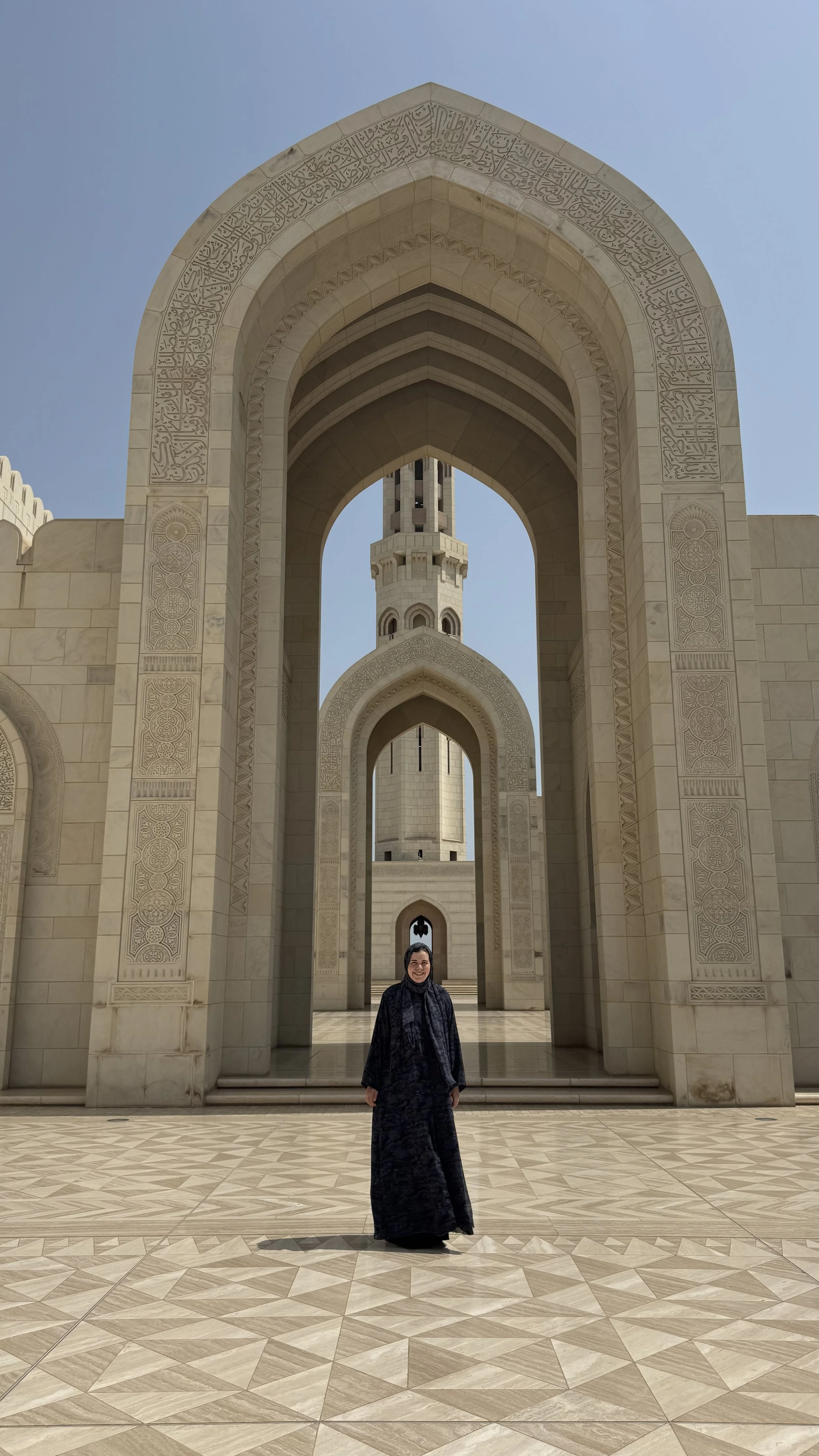 Eine Frau in schwarzem Kleid steht vor einer großen Architektur mit islamischen Elementen, darunter große Bögen und einen Minarett-Turm im Hintergrund, aus rohem Marmor gebaut.