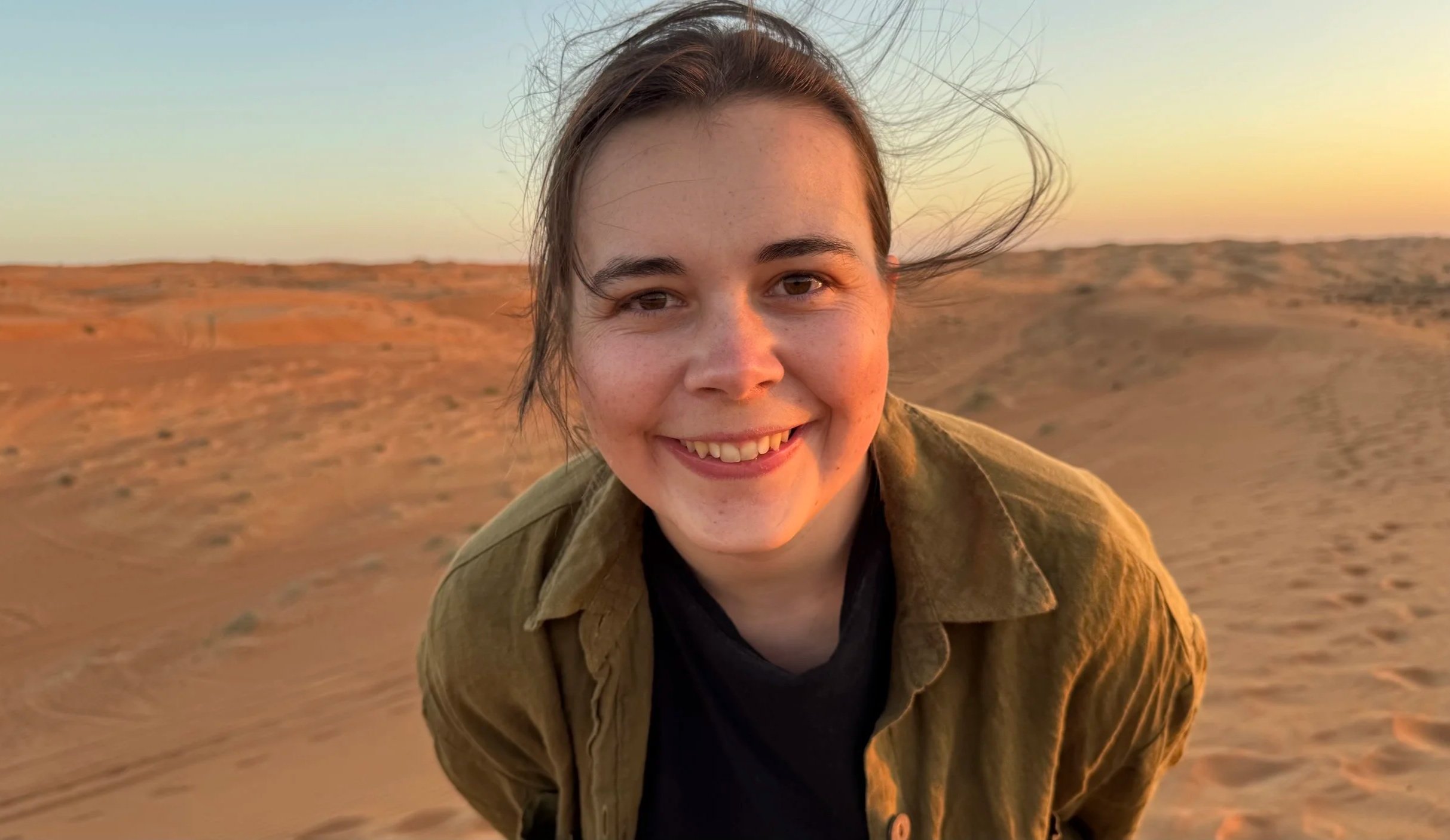 Junge Frau lächelt in einer Wüstenlandschaft bei Sonnenuntergang, mit windzerzausten Haaren und leicht zerzaustem Hemd.