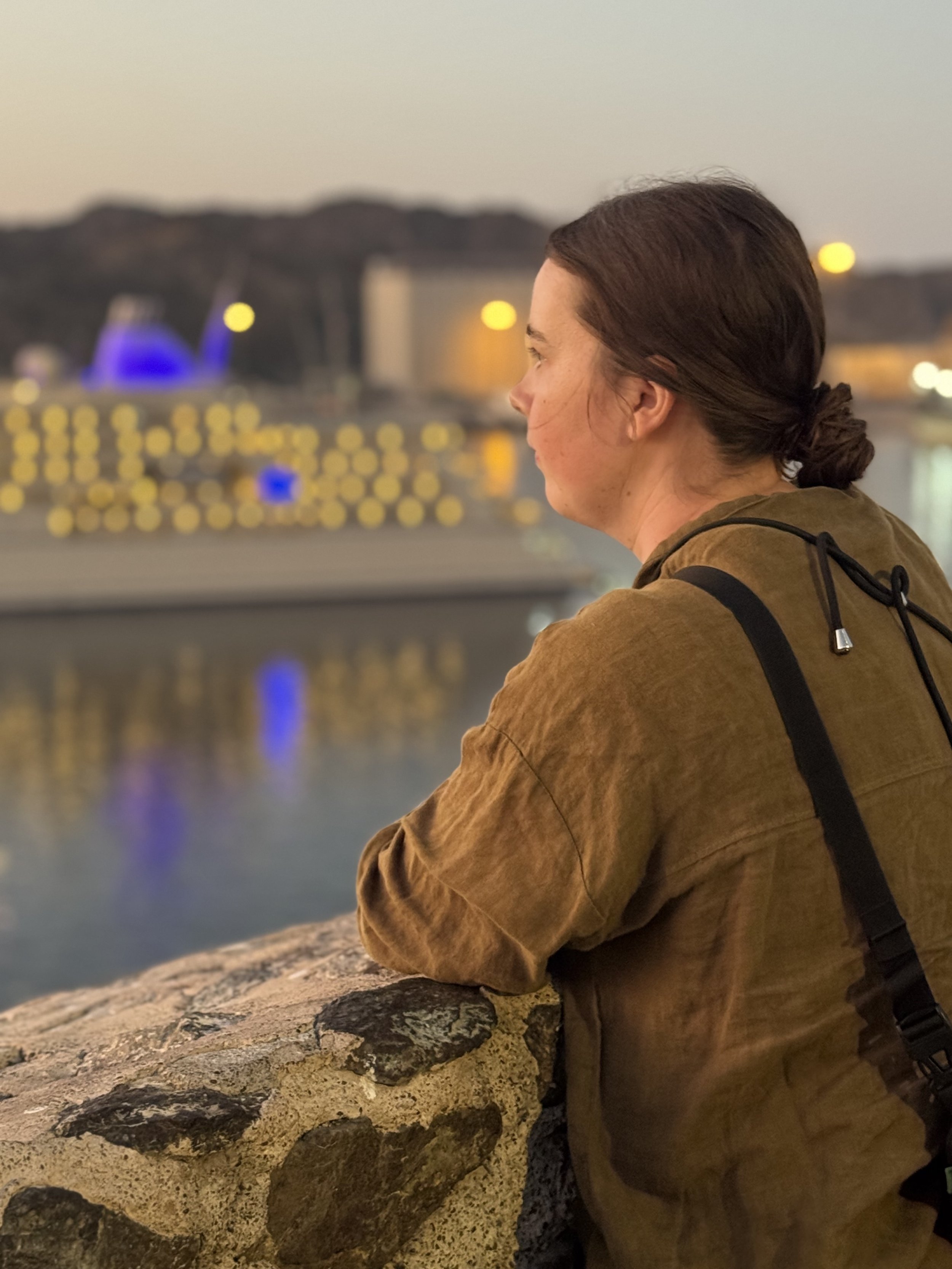 Junge Frau mit braunen Haaren im Profil, sitzt an einem Steinkamin am Wasser bei Sonnenuntergang, im Hintergrund eine Brücke und Lichter, ruhige Atmosphäre.