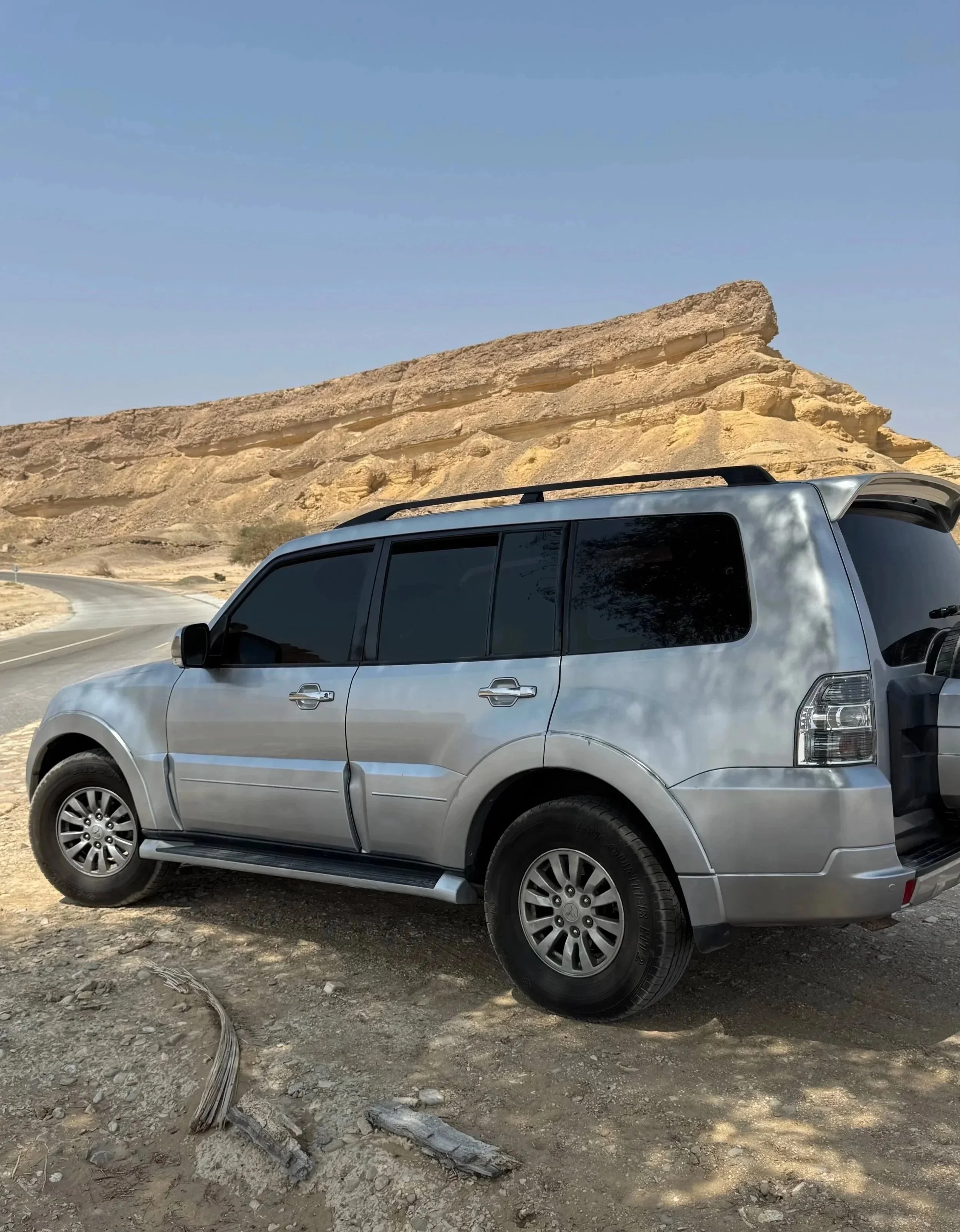 Ein silberner SUV steht auf unbefestigtem, staubigem Boden neben einer Wüstenlandschaft mit großen Sandsteinformationen. Der Himmel ist klar und blau.