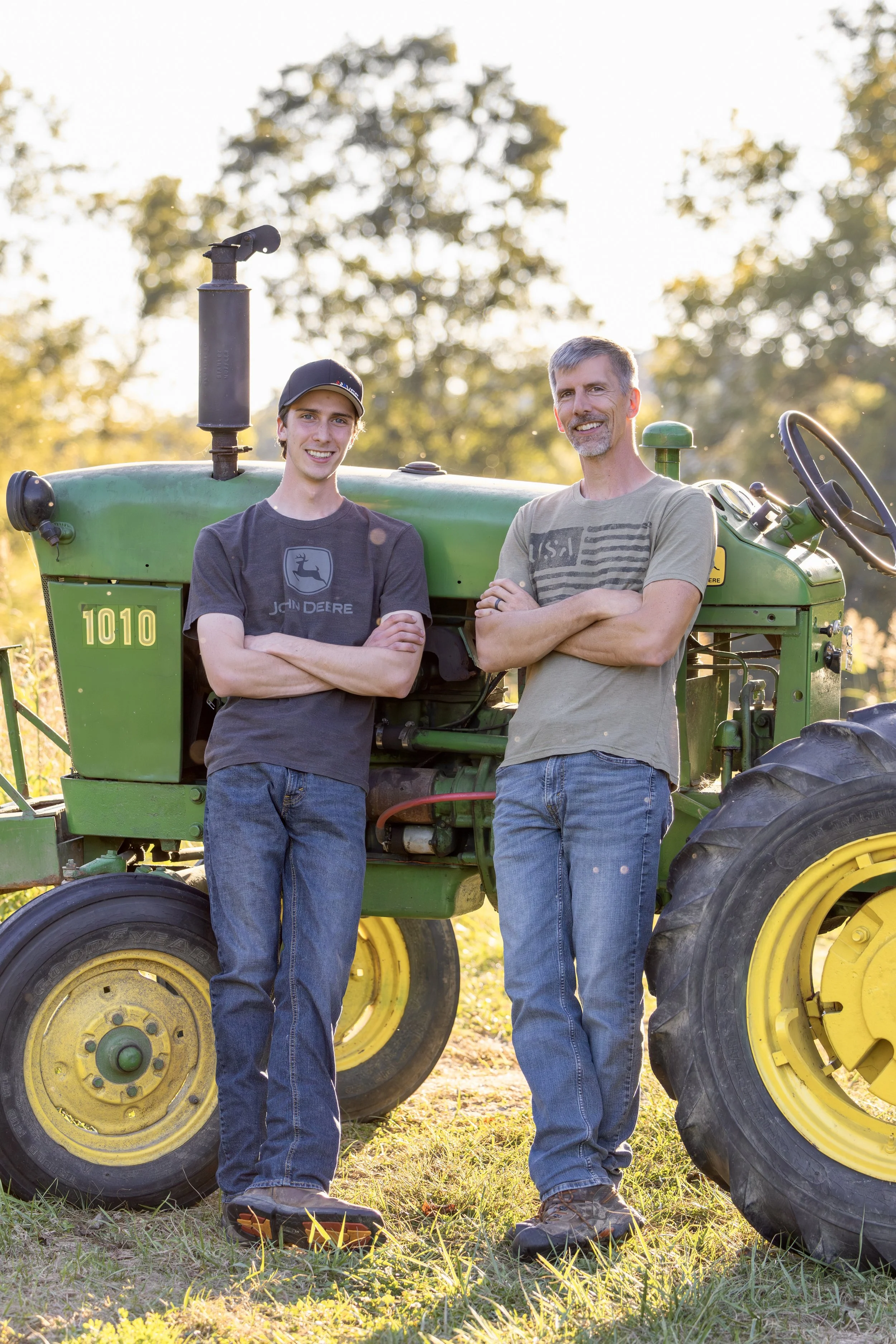 Two men standing beside a green John Deere tractor outdoors in the late afternoon, smiling and posing with arms crossed, on a grassy field with trees in the background.