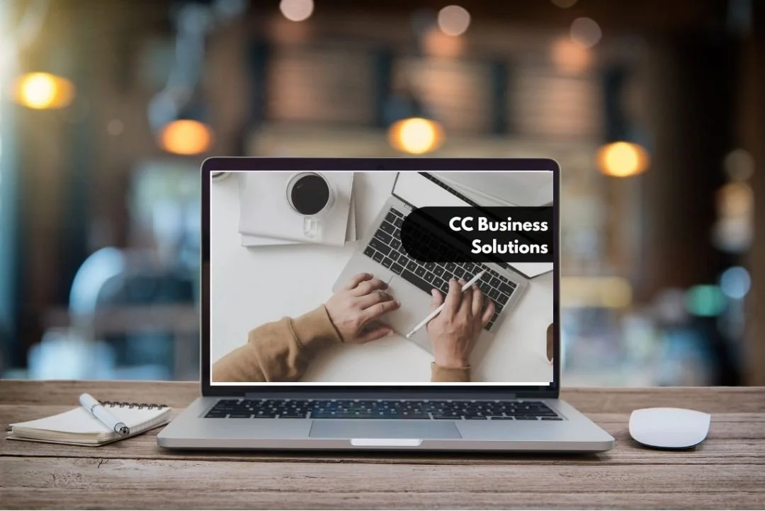 Laptop on a wooden desk displaying a presentation slide titled 'CC Business Solutions' with a hand using a stylus on a keyboard, a cup of coffee, a notepad with a pen, and a computer mouse in a blurred background.