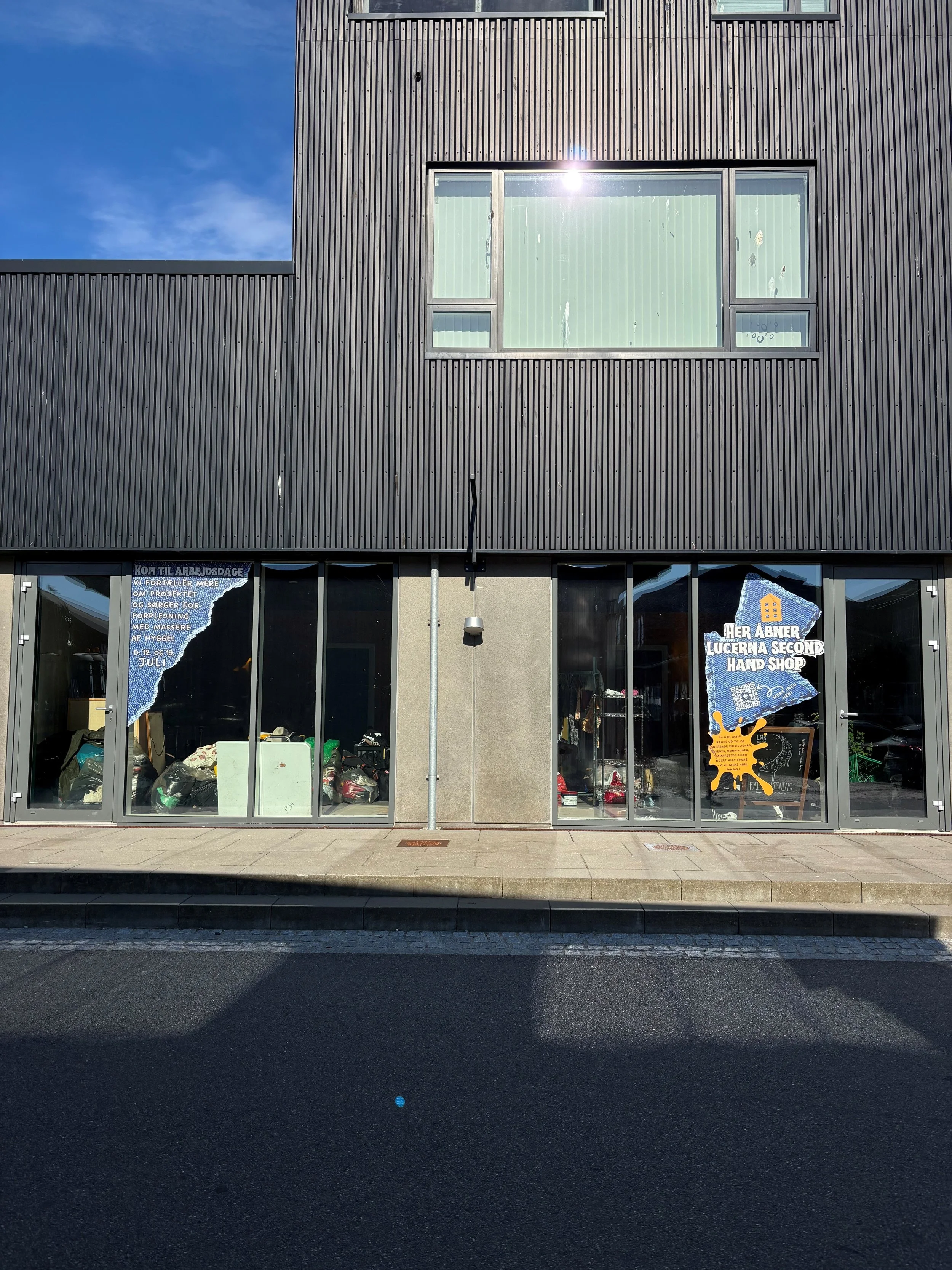Butiksvindue med glasdøre, hvor der er plakater. Plakaten til venstre er delvist revet i stykker og har tekst om arbejdsdage. Plakaten til højre er reklame for Lucerna Second Hand Shop, med en gul icon og tekst på dansk.