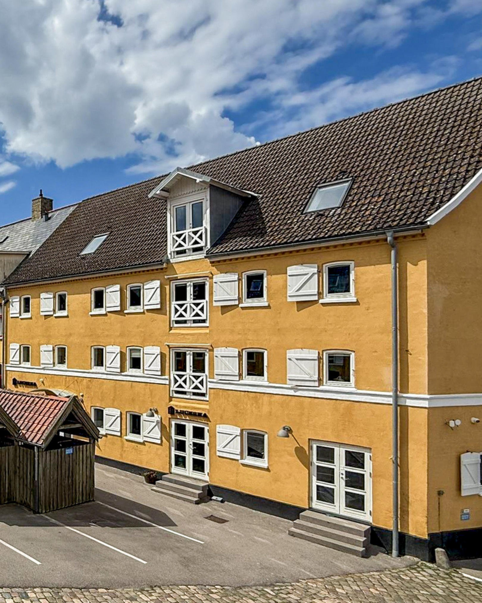 Et gult, fire etager højt bygning med hvide vinduesrammer, hvide udhæng, en balkon i midten og en tagterrasse under skråtag med tagvinduer. Der er en parkeringsplads foran bygningen.