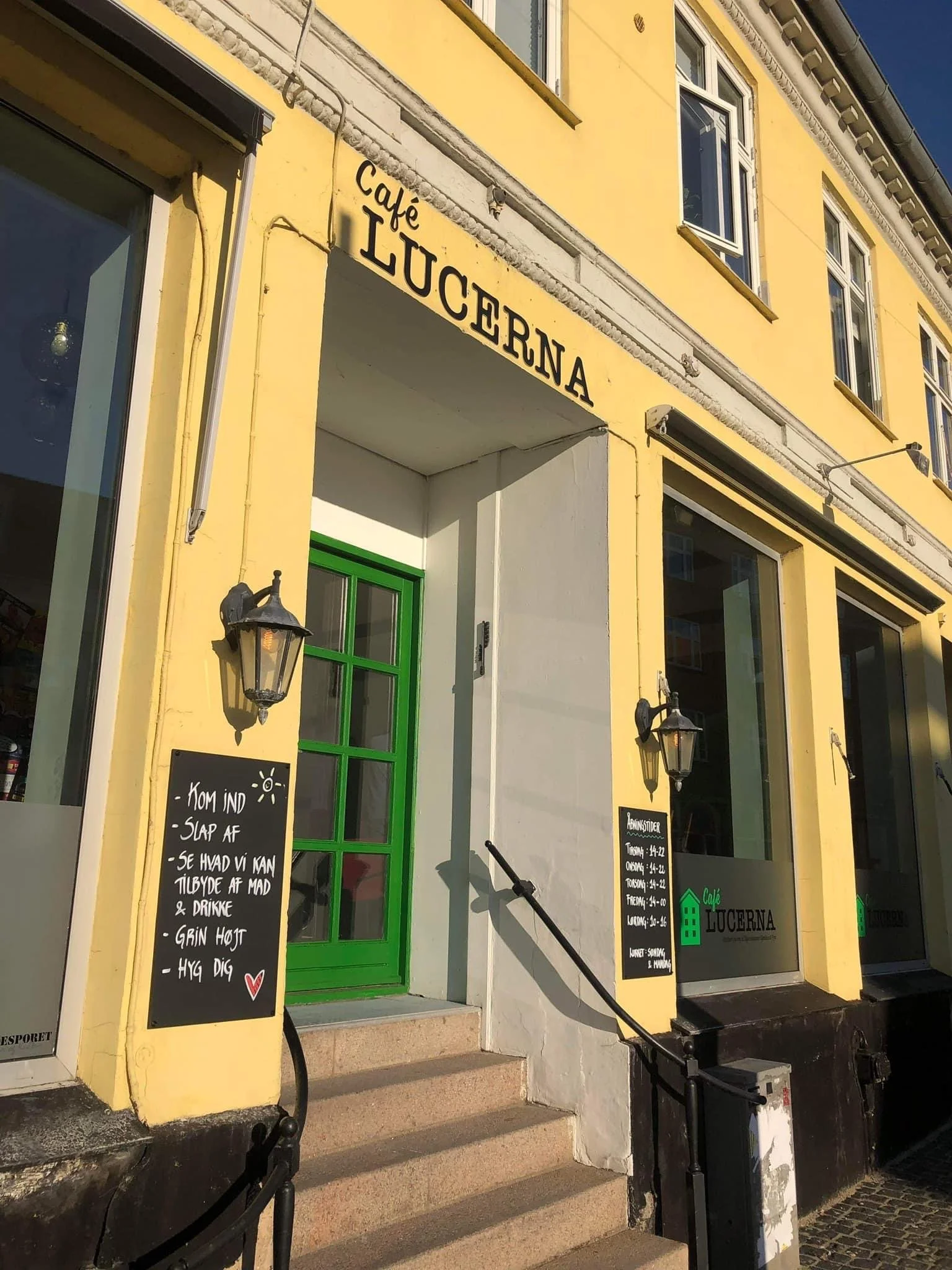 Billedet viser en café kaldet 'Café Lucerna' udenfor, med gardiner, vinduer og skilte med åbningstider og tilbud. Der er trapper og sten på fronten.