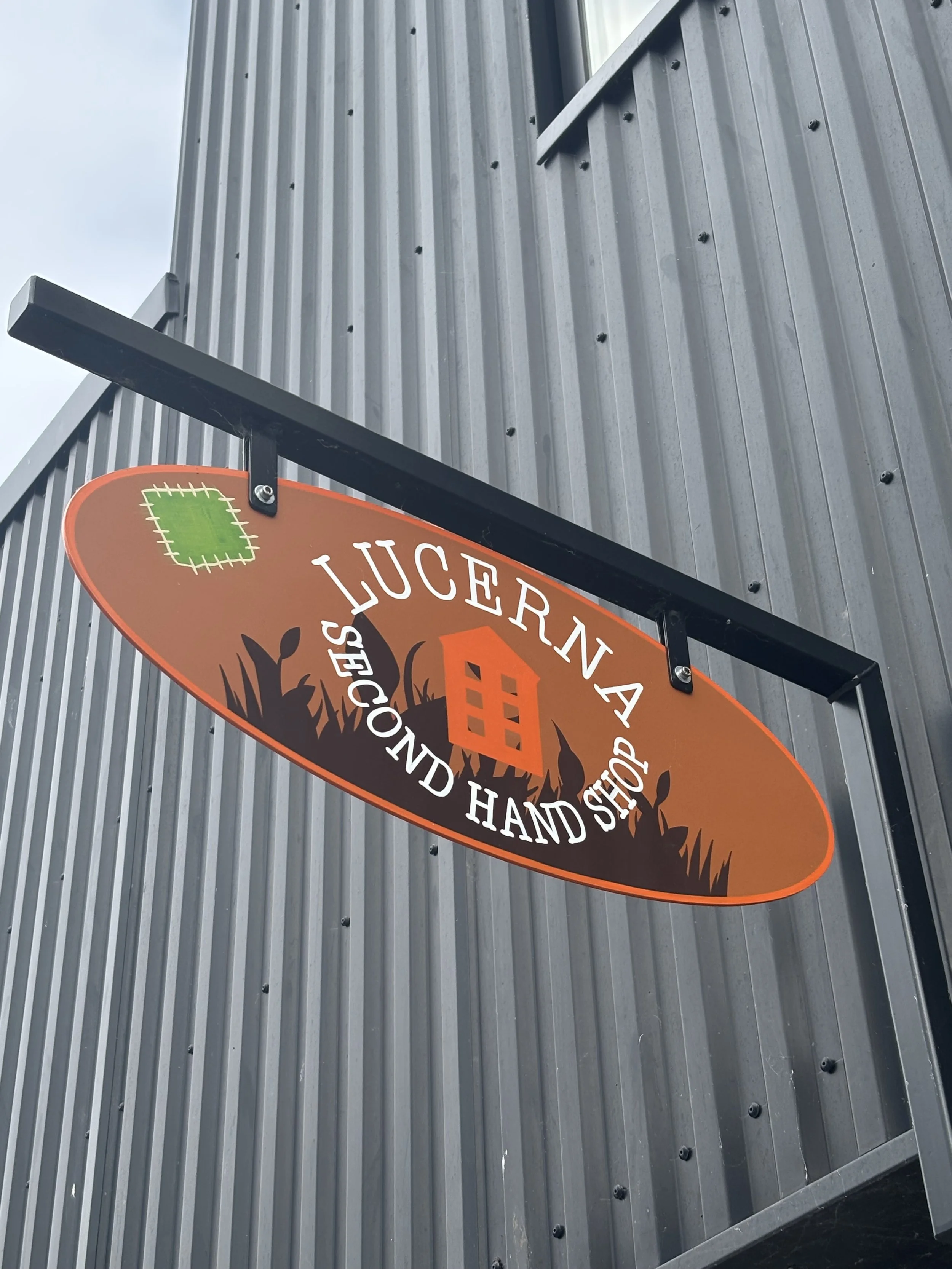 Skilt på en bygning med teksten 'LUCERNA' og 'SECOND HAND SHOP' med en illustration af et hus og græs, monteret på en metalvæg.
