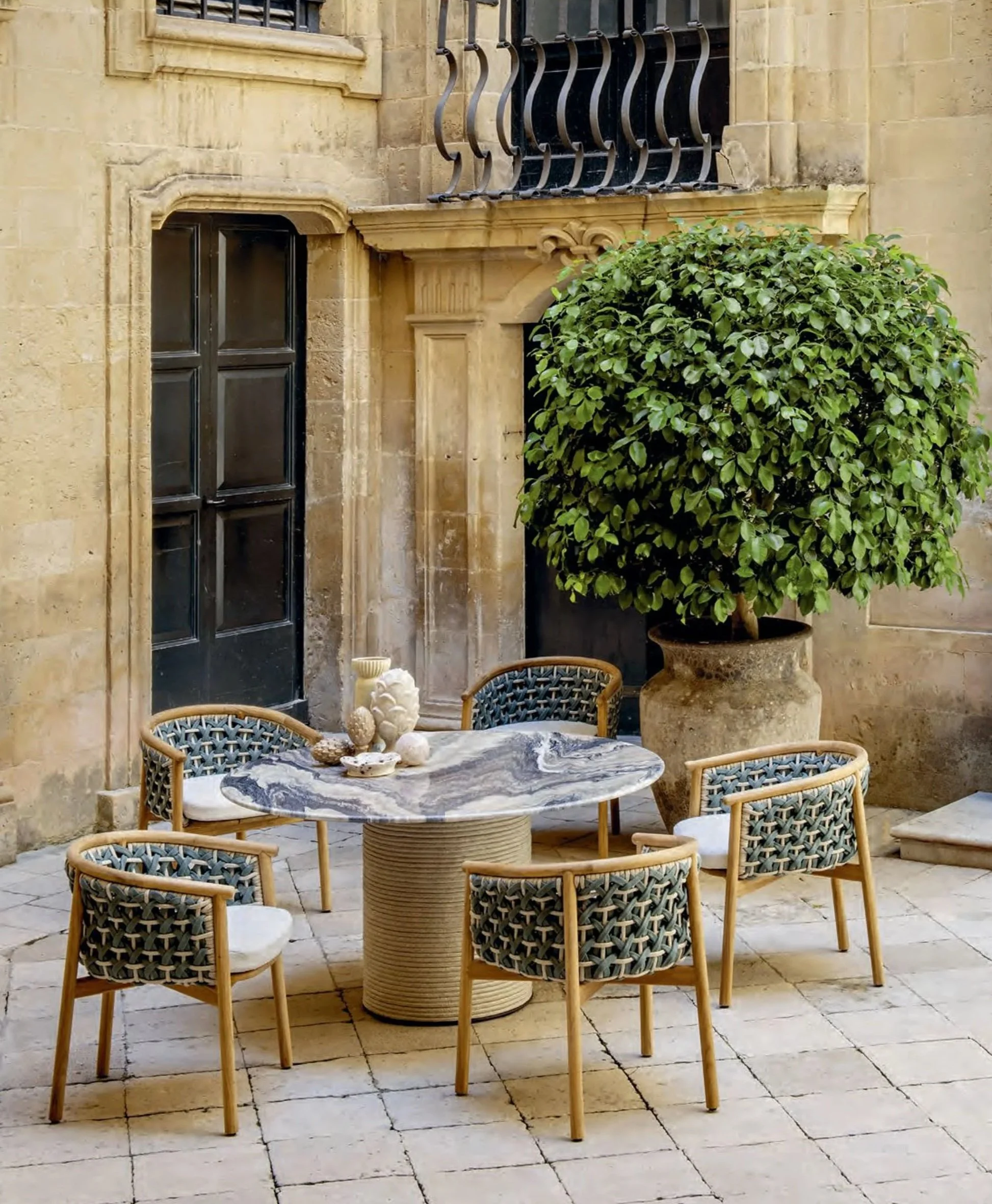 An outdoor courtyard with a round marble table, wicker chairs, a large potted shrub, decorative stone wall, and small sculptures on the table.